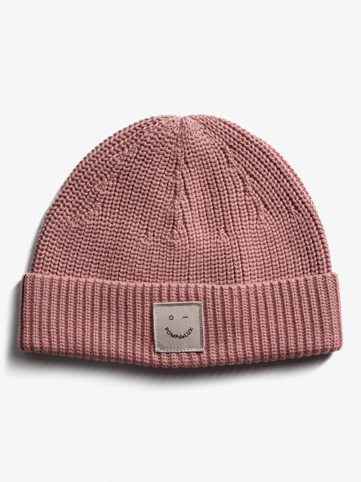 Eine rosafarbene Strickmütze mit umgeschlagenem Rand zeigt ein Quadratlabel mit einem minimalistischen Gesicht und dem Text "POMPdeLUX" auf weißem Hintergrund.