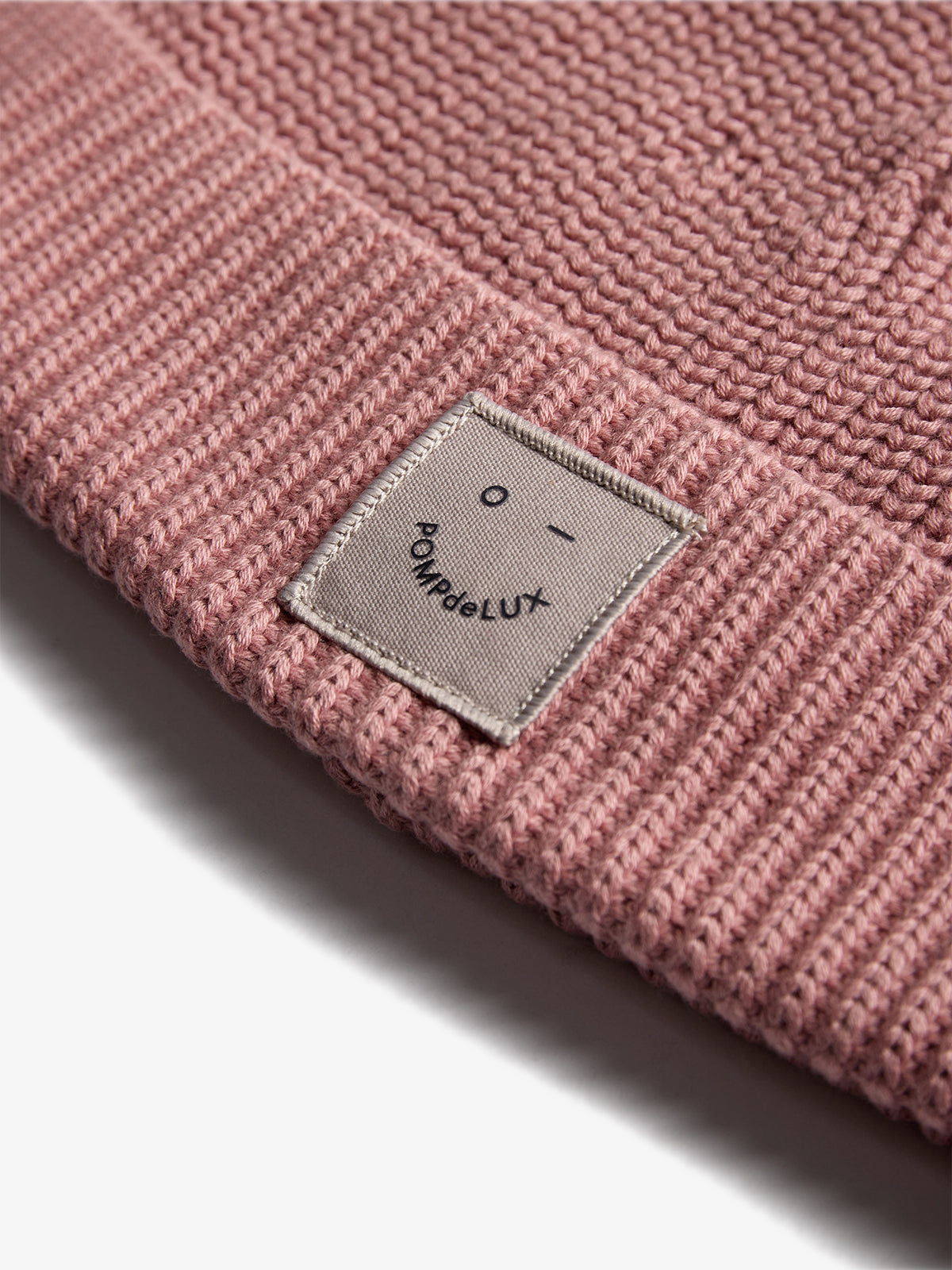 Eine rosafarbene Strickmütze liegt auf einer Oberfläche. Ein aufgenähtes Etikett zeigt ein einfaches Gesicht und den Text "Pomp de Lux". Sie hat eine dezente Struktur.