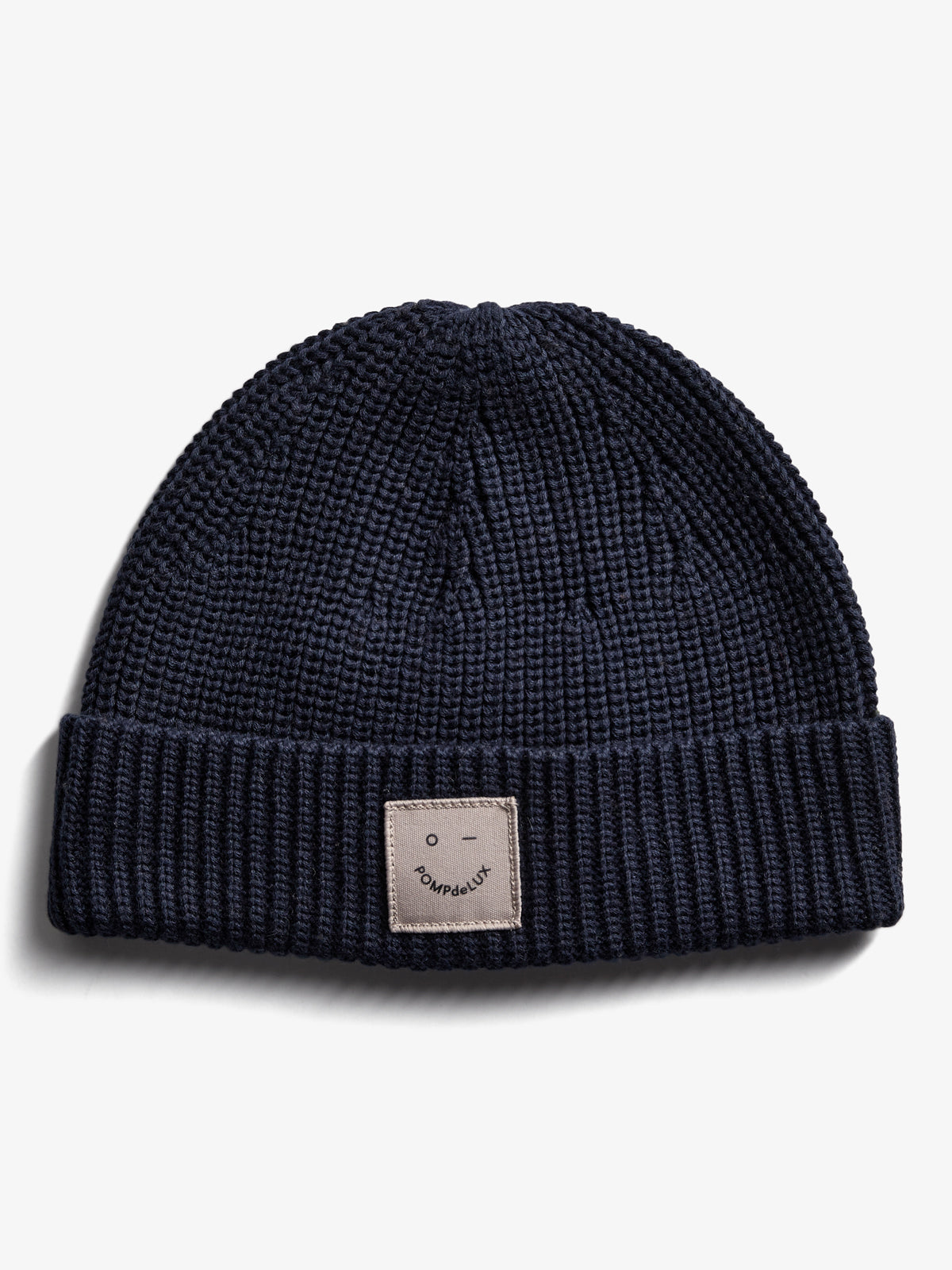 Eine dunkelblaue Strickmütze liegt auf einer weißen Fläche. Auf der Vorderseite befindet sich ein rechteckiges Etikett mit der Aufschrift „POMPdeLUX“ und einem minimalistischen Gesichtssymbol.