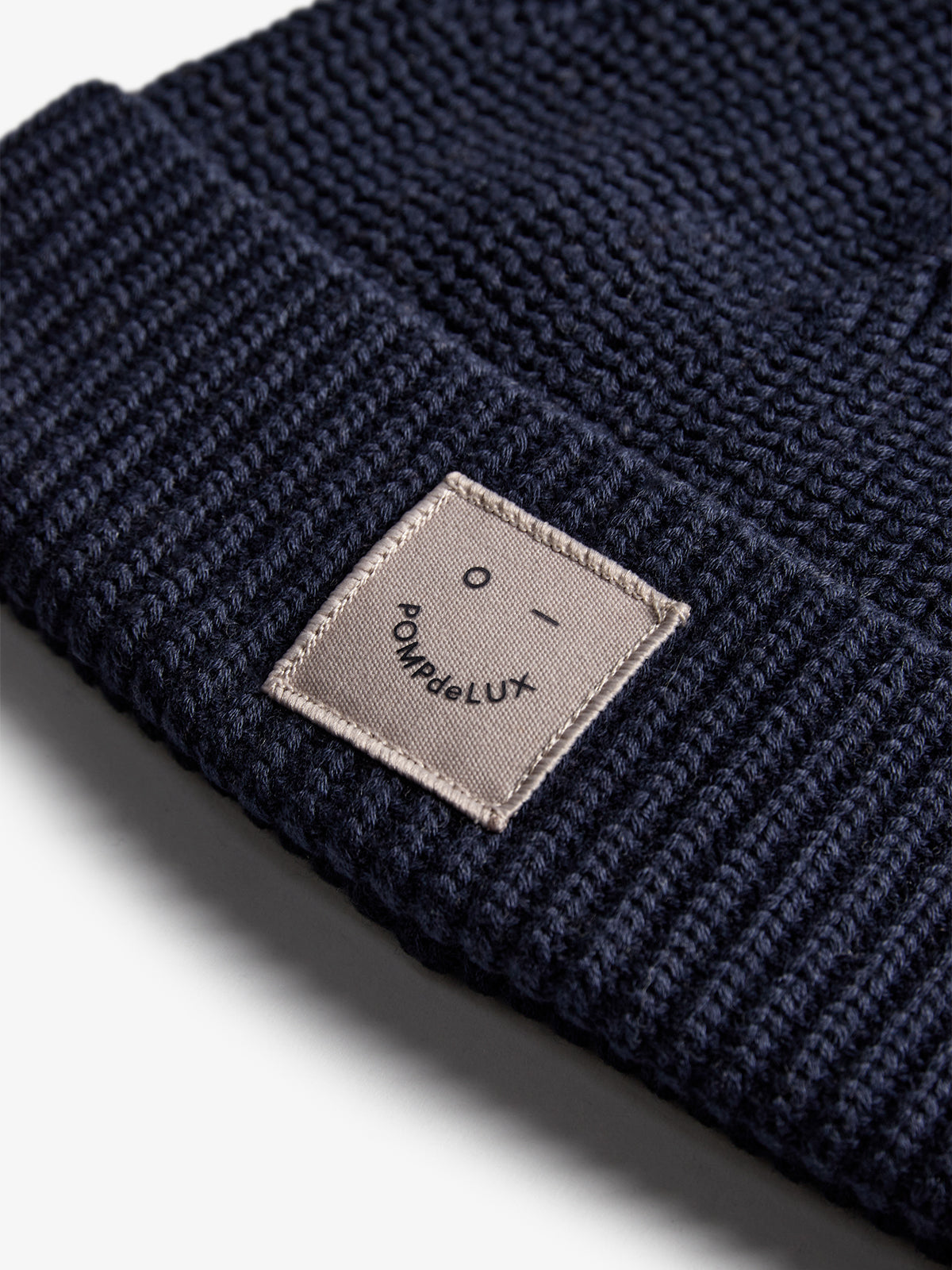 Eine dunkelblaue Strickmütze zeigt vorne ein rechteckiges Etikett mit der Aufschrift "POMPdeLUX" und einem minimalistischen Gesichtssymbol auf weißem Hintergrund.
