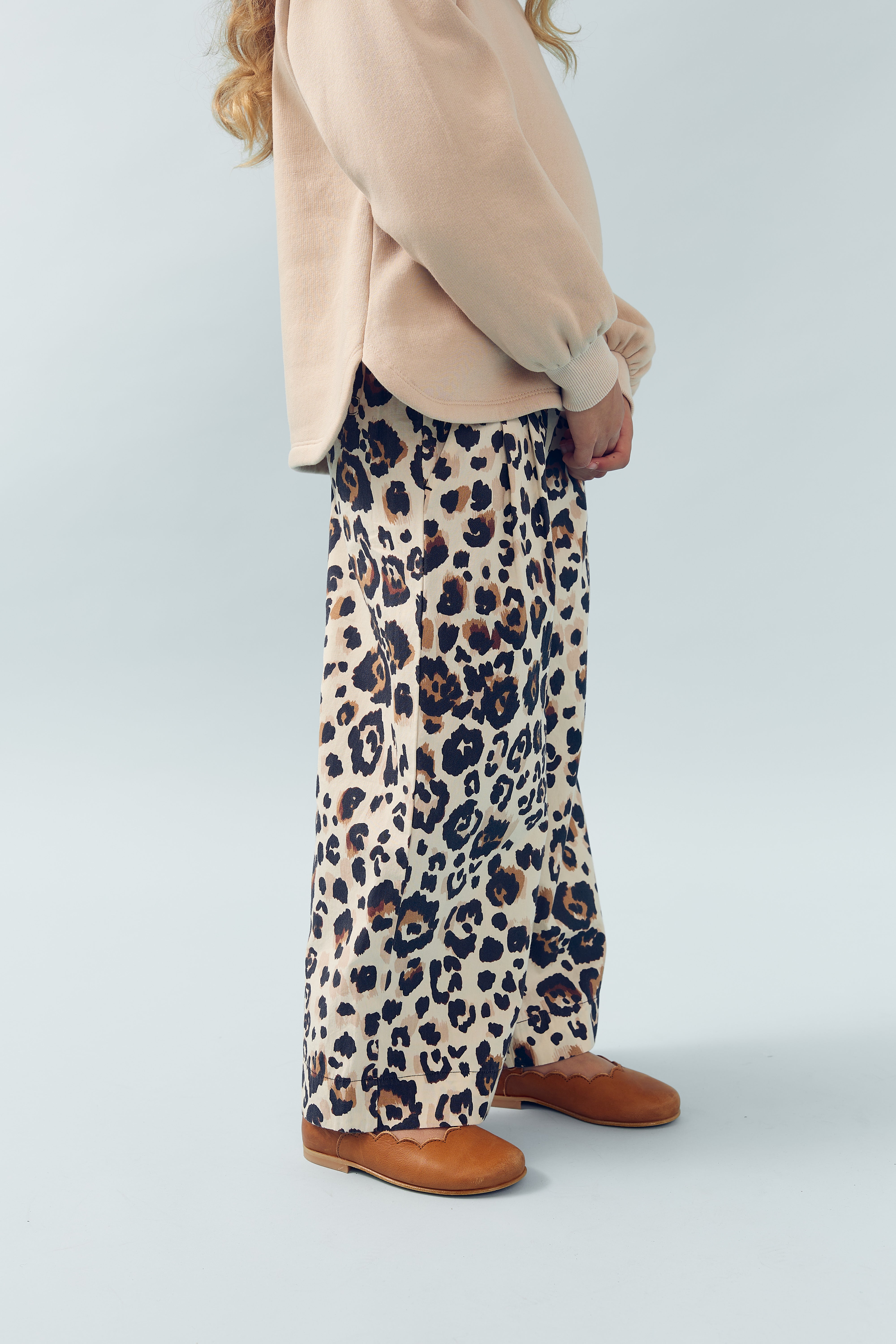 Ein Kind trägt eine beige Bluse und weite, leopardenmusternde Hosen. Es steht ruhig auf einem hellen Hintergrund, trägt braune Schuhe und die Haare sind lang und lockig.