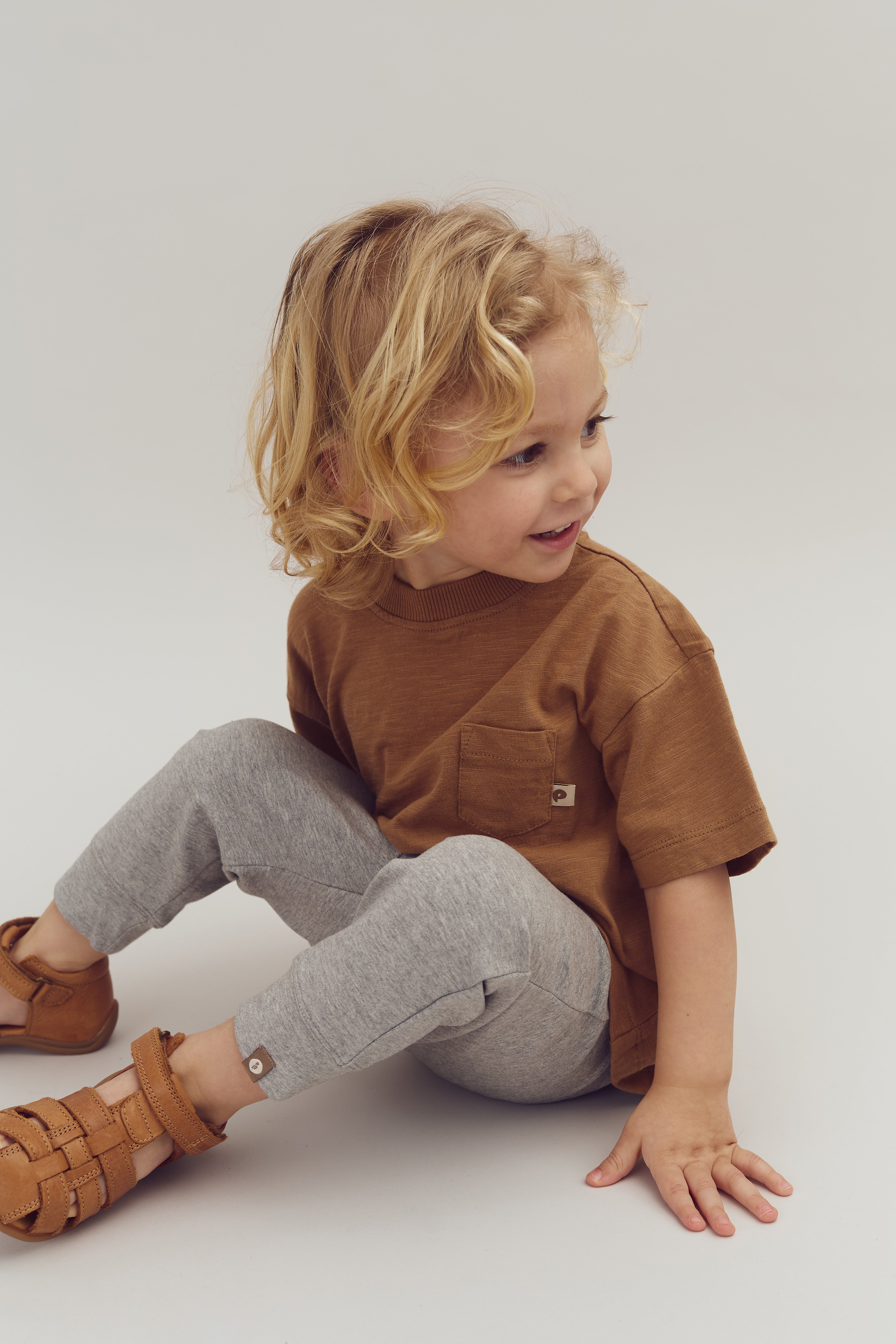 Ein kleines Kind sitzt lächelnd auf dem Boden, trägt ein braunes T-Shirt, graue Leggings und braune Sandalen. Der Hintergrund ist schlicht und einfarbig.