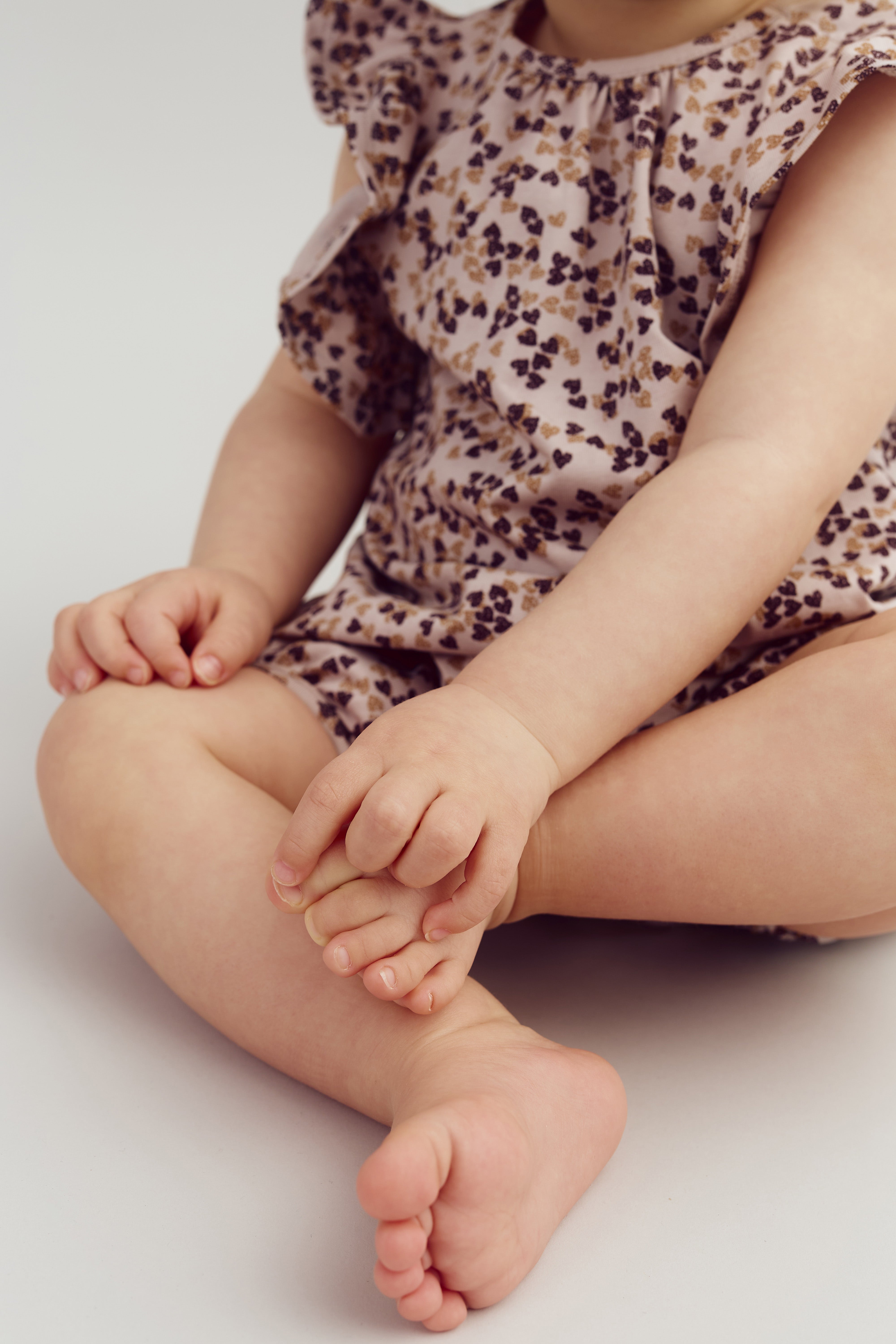 Kind sitzt mit gekreuzten Beinen auf hellem Boden, trägt ein geblümtes Kleid und hält einen Fuß mit den Händen. Der Hintergrund ist neutral gestaltet.