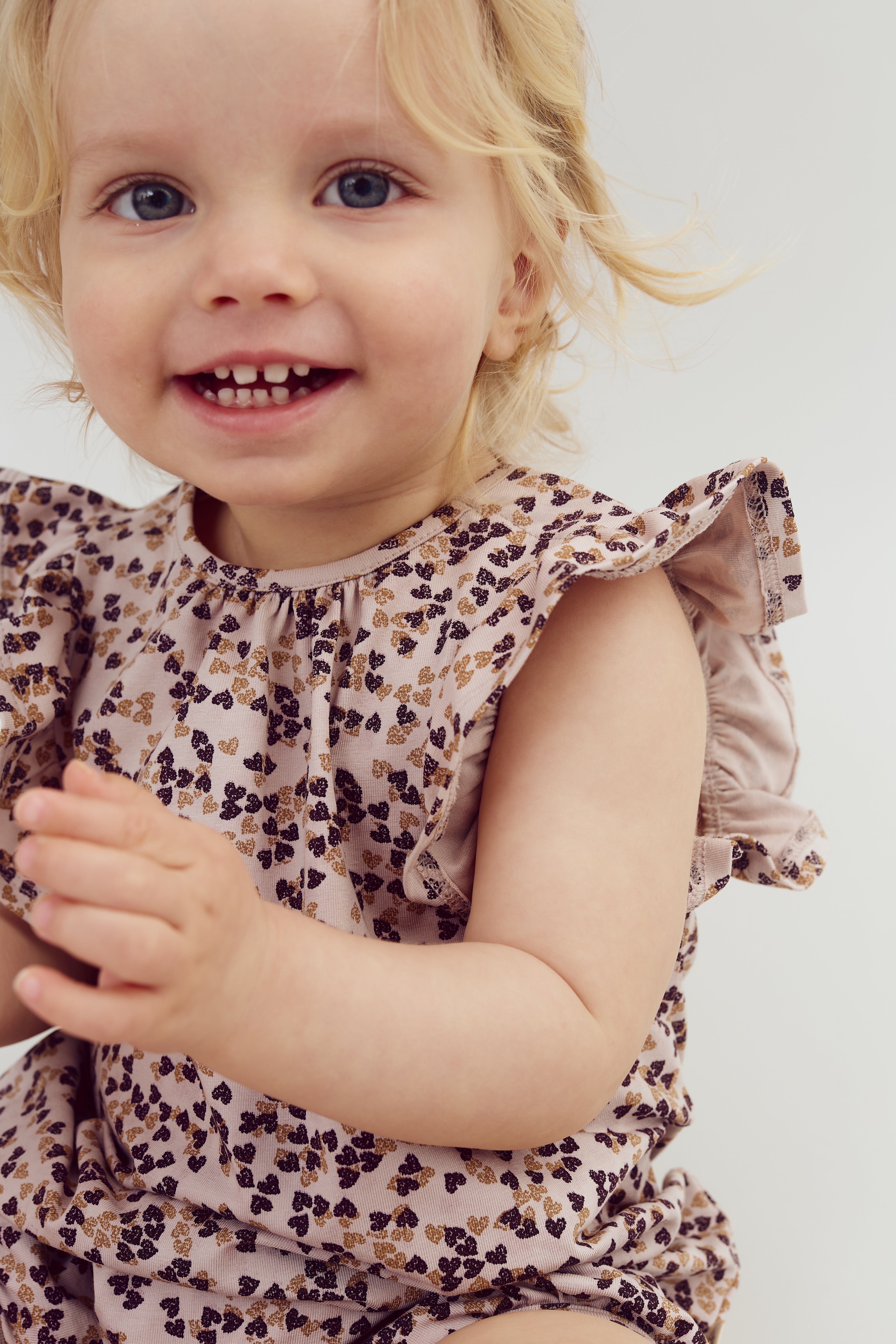 Ein kleines Kind, das lachend in die Kamera schaut, trägt ein ärmelloses Kleid mit blumigem Muster. Der Hintergrund ist schlicht und weiß.