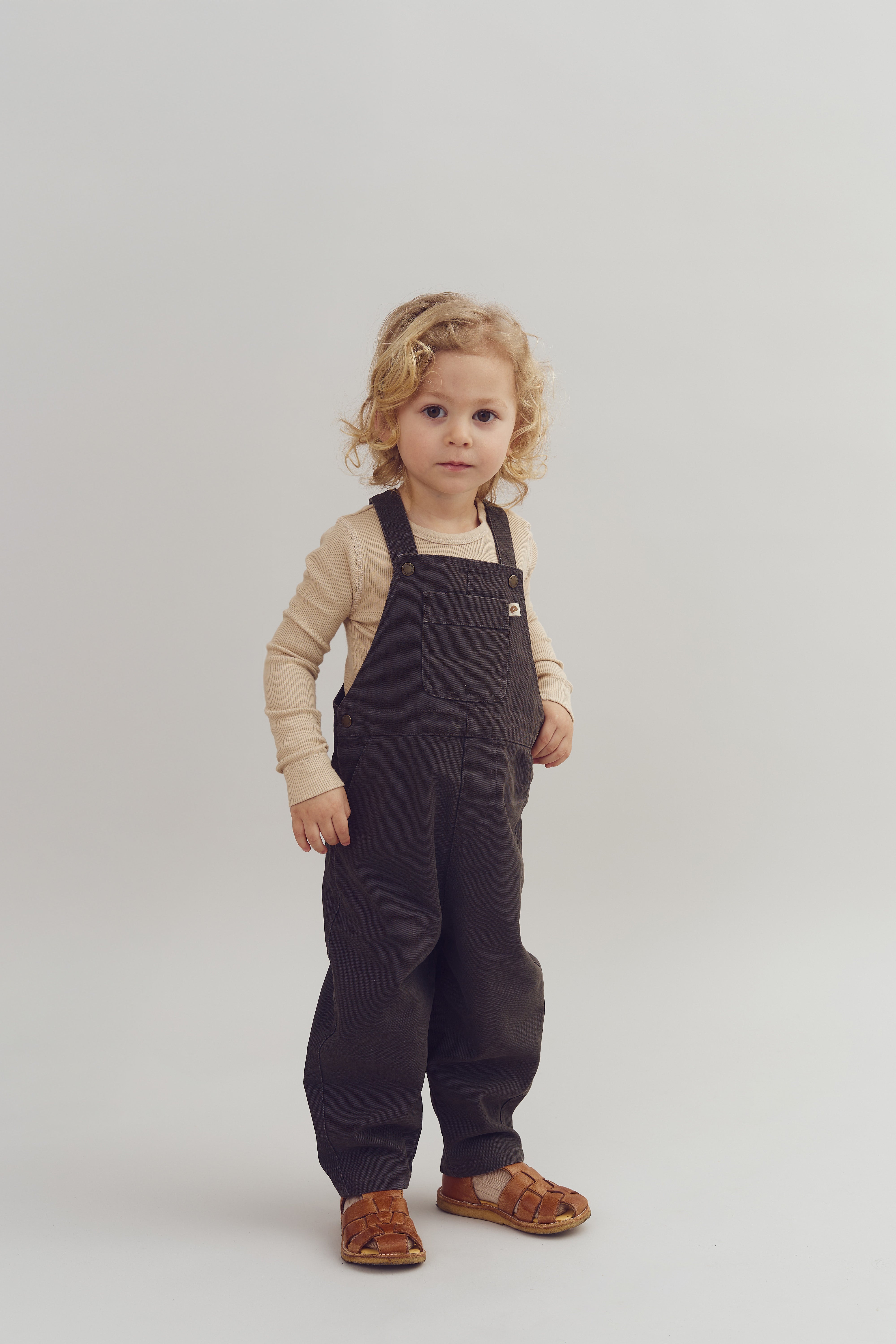 Ein Kind steht still, trägt eine beigefarbene Langarmbluse und eine braune Latzhose. Es hat lockiges blondes Haar und braune Sandalen, auf weißem Hintergrund.