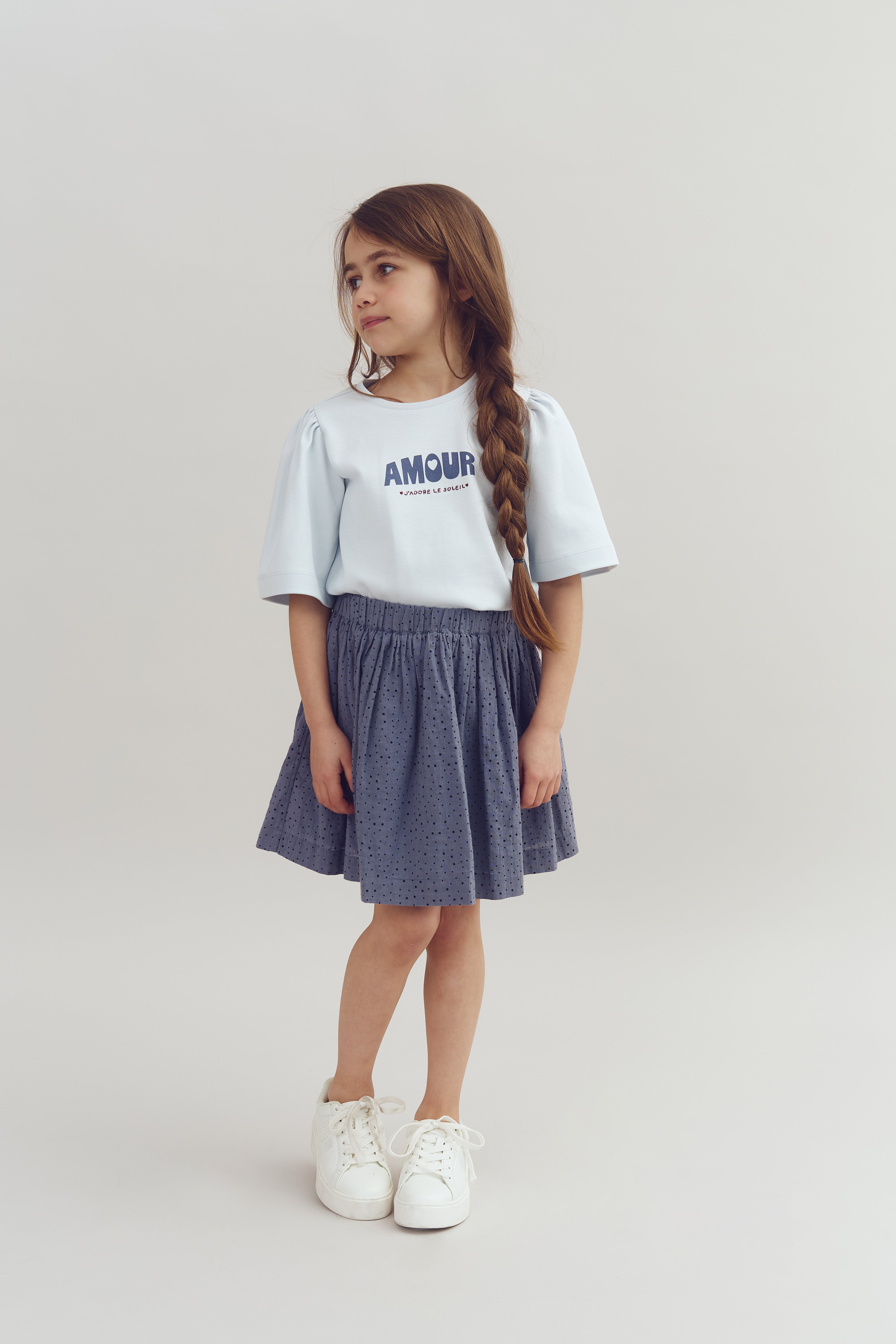 Ein Mädchen steht in einem hellen Raum. Sie trägt ein weißes T-Shirt mit der Aufschrift "AMOUR" und einen blau gepunkteten Rock. Sie schaut erwartungsvoll zur Seite.