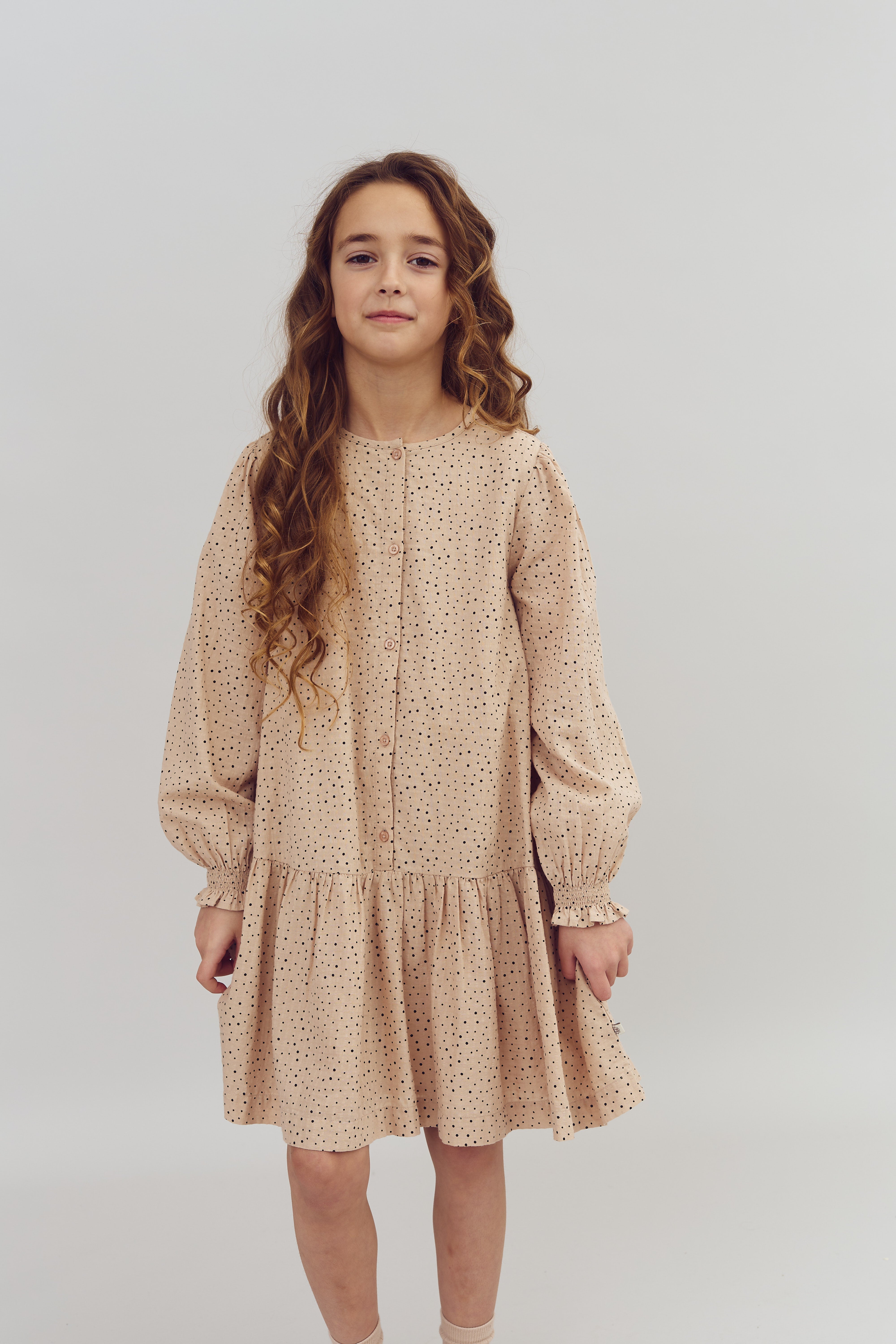 Mädchen trägt ein gepunktetes, pfirsichfarbenes Kleid, steht ruhig. Ihr Haar ist lang und wellig. Der Hintergrund ist einfarbig und neutral, was das Motiv hervorhebt.
