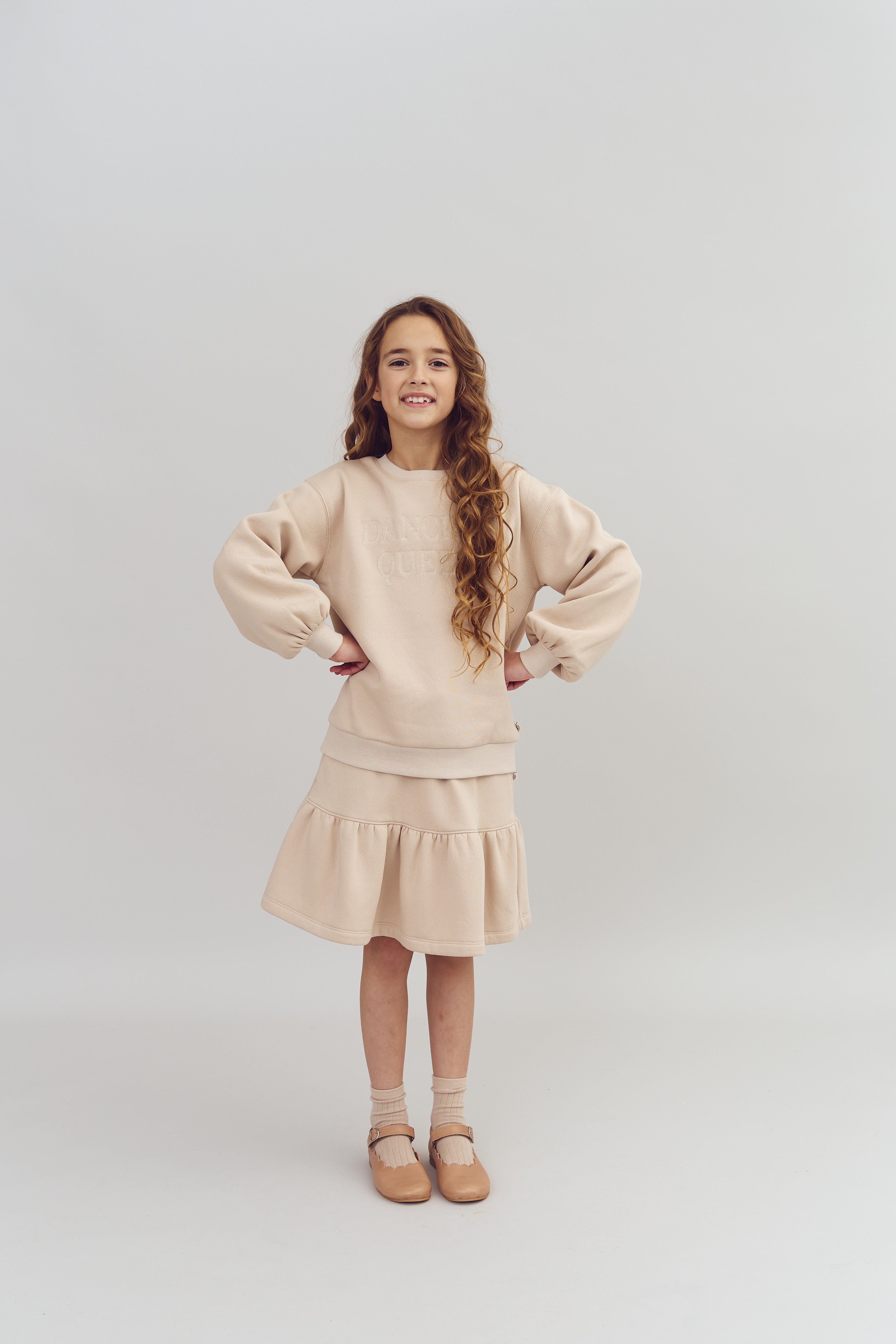 Mädchen steht mit den Händen in der Hüfte, trägt beige Kleidung mit der Aufschrift "Dance Queen", in einem neutralen Studiohintergrund. Sie lächelt und trägt braune Schuhe.