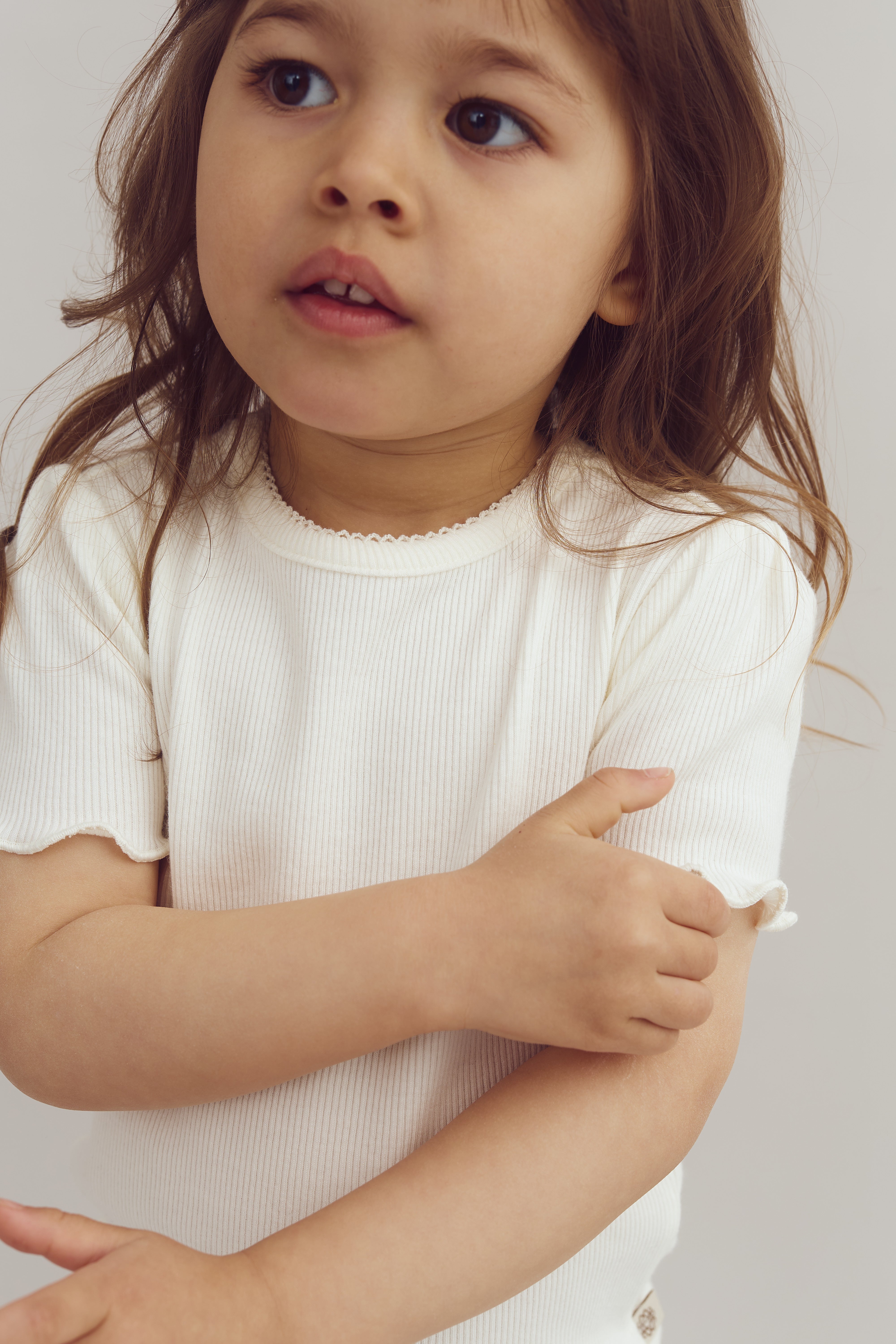Ein kleines Kind trägt ein cremefarbenes, geripptes T-Shirt und verschränkt die Arme vor der Brust. Der Hintergrund ist neutral und schlicht.
