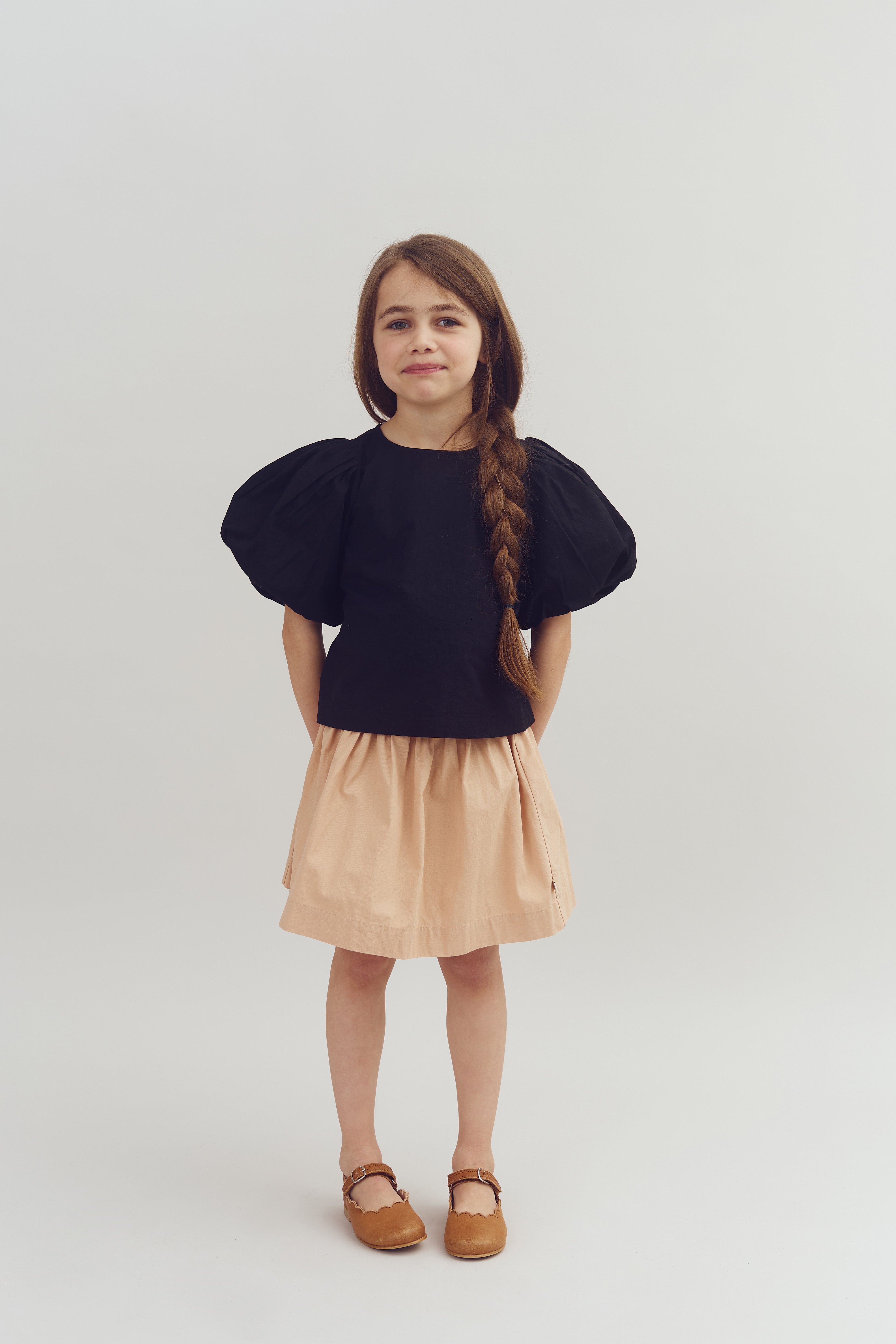 A young girl stands against a plain background, wearing a black top with puffed sleeves, a beige skirt, and brown shoes. Her hair is styled in a long braid.