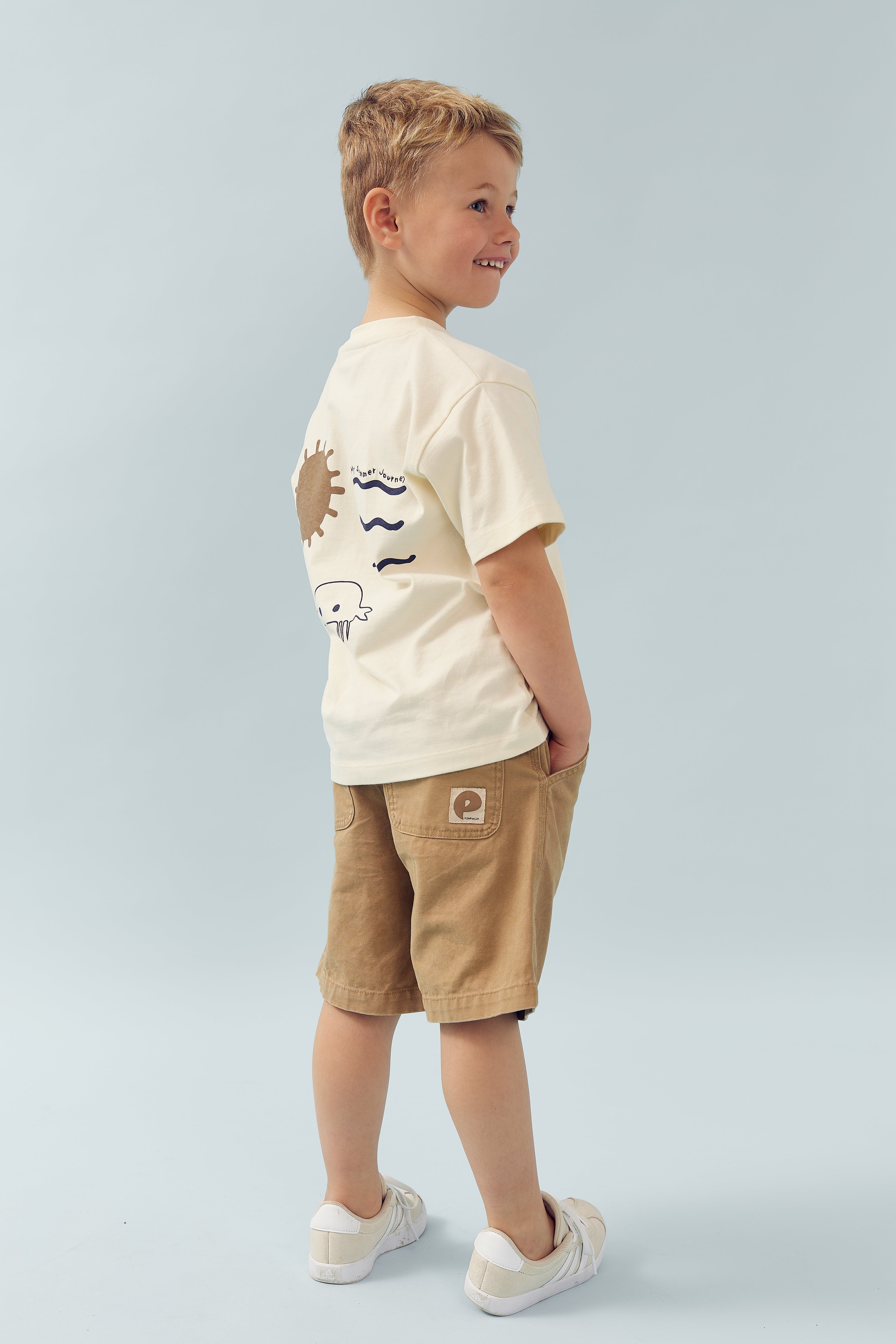 Ein Junge trägt ein cremefarbenes T-Shirt mit Sonnen- und Wellenmotiven, braune Shorts und weiße Turnschuhe. Er steht lächelnd auf hellem Hintergrund mit den Händen in den Taschen.