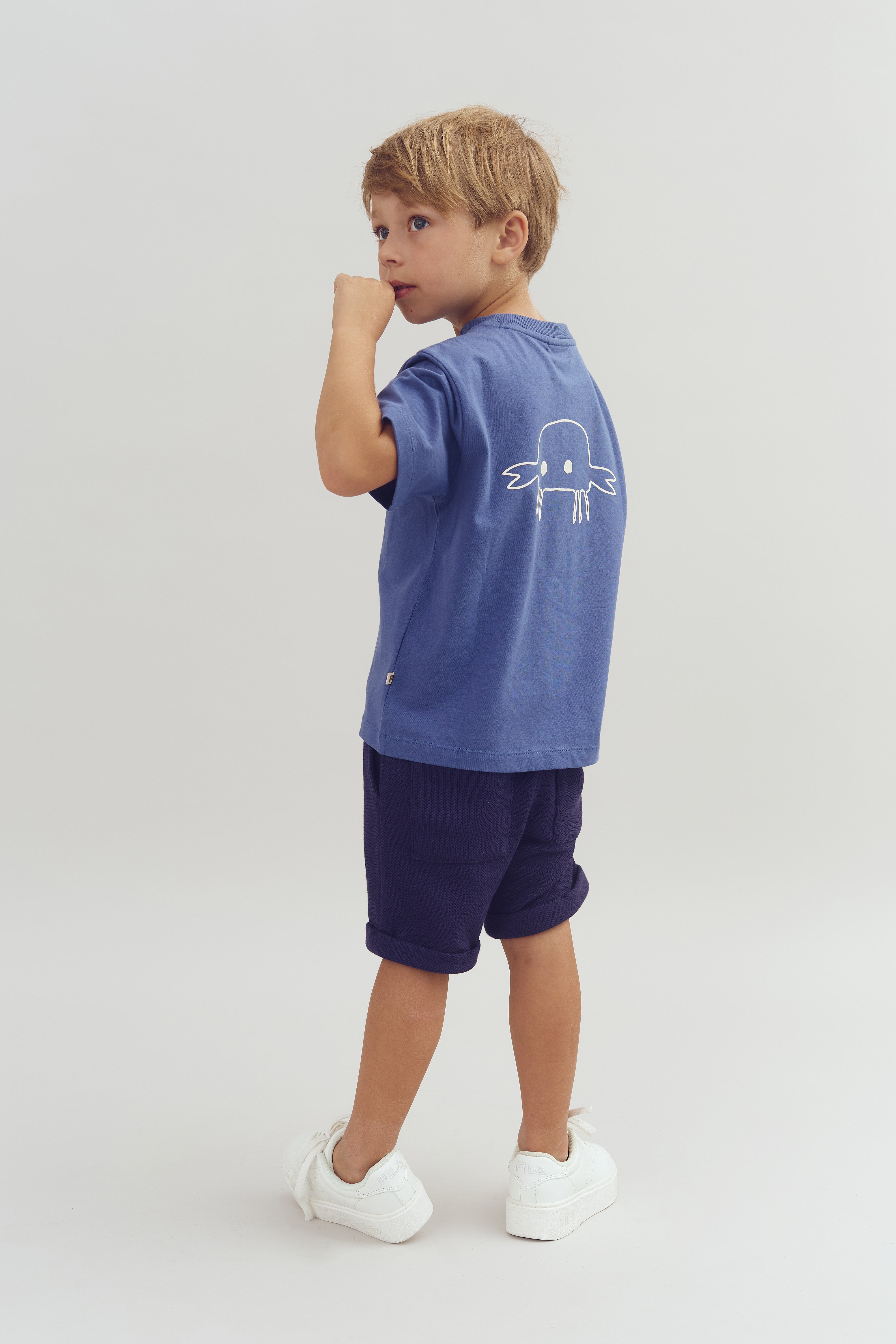 Ein Junge trägt ein blaues T-Shirt mit einem weißen Oktopusmotiv auf der Rückseite, dazu dunkelblaue Shorts und weiße Turnschuhe. Er blickt zur Seite in einem neutralen, hellen Raum.