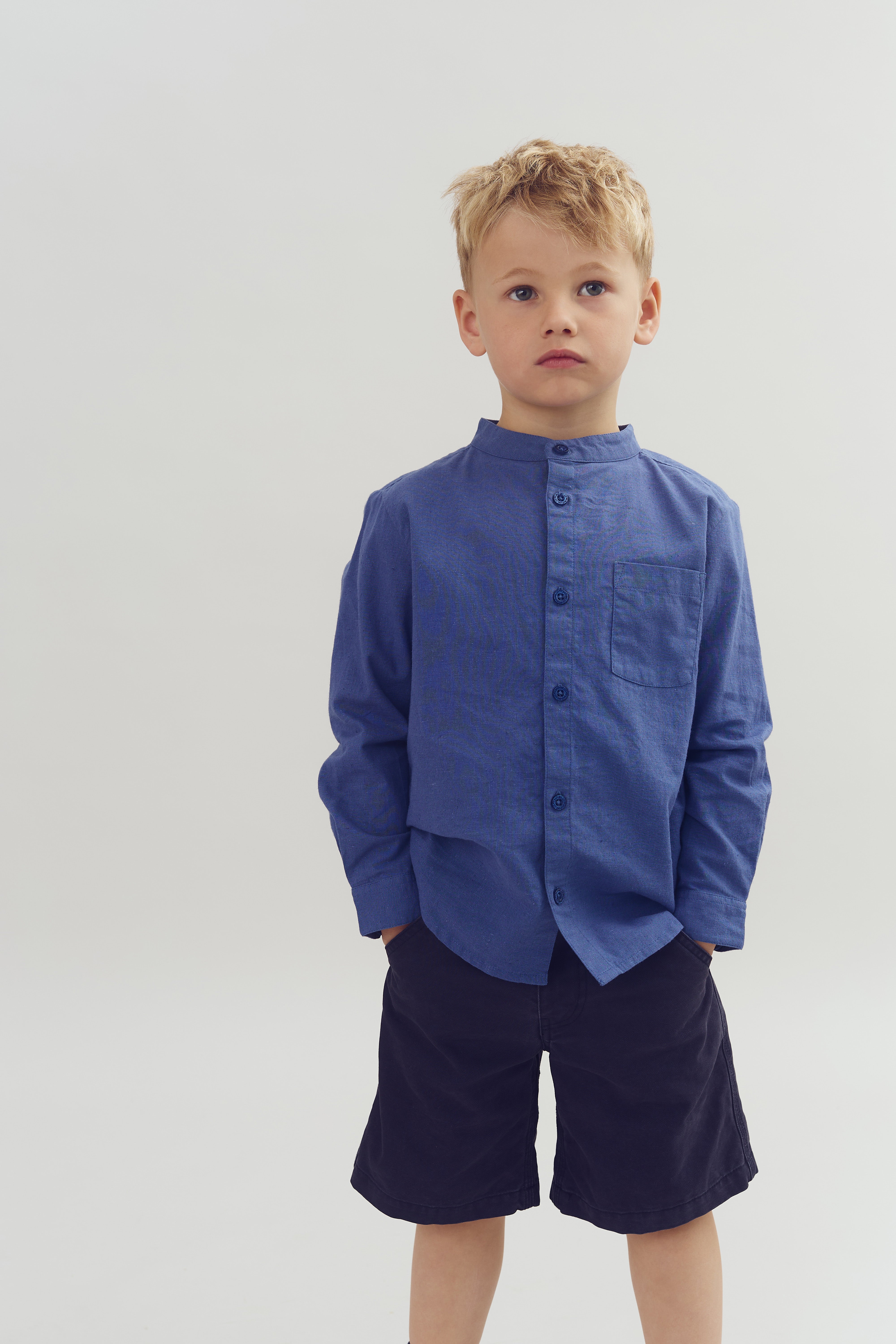 Ein Junge trägt ein blaues Hemd und schwarze Shorts. Er steht ruhig mit den Händen in den Taschen vor einem einfachen, grauen Hintergrund.
