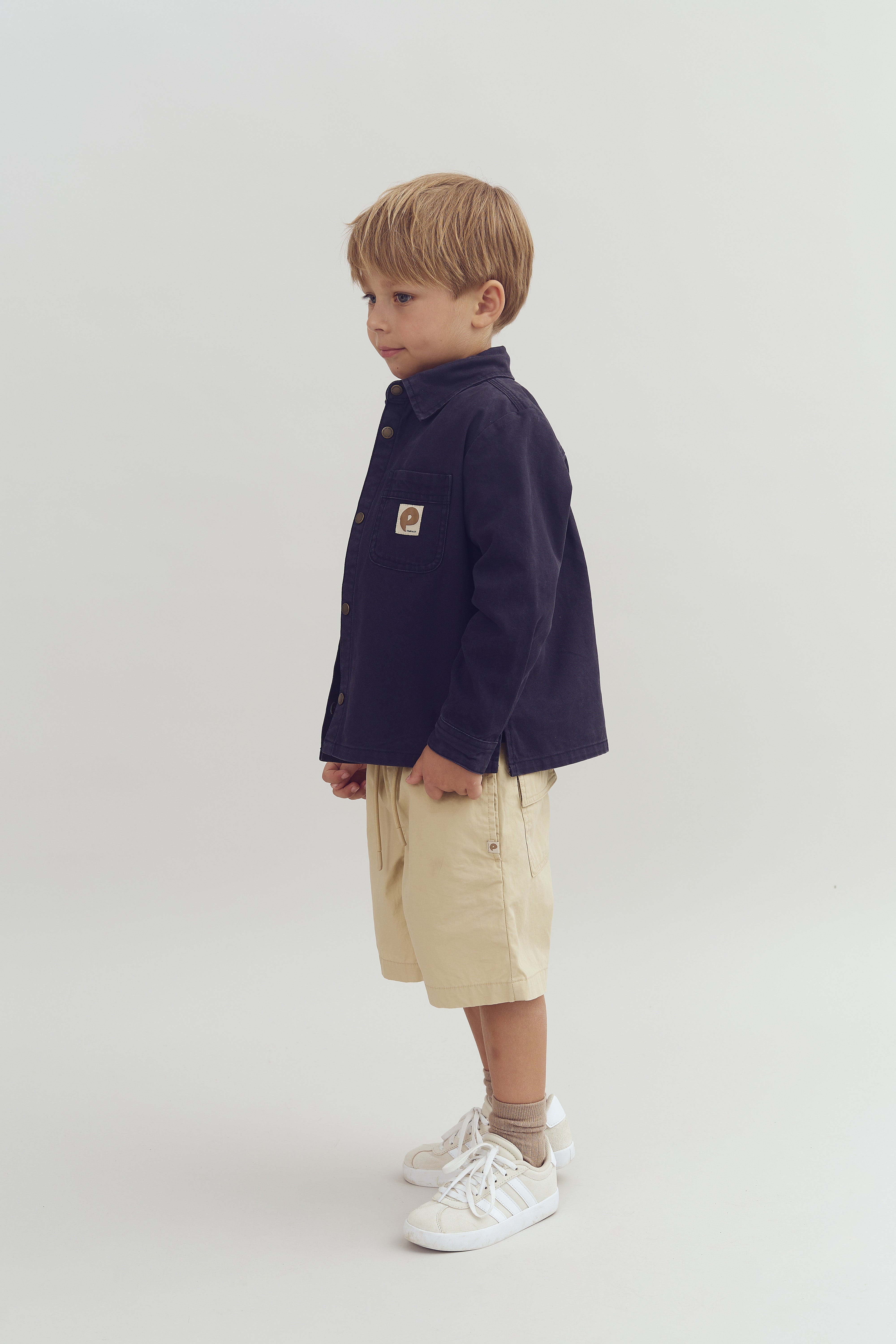 Ein Junge steht seitlich, trägt eine dunkelblaue Jacke mit einem Logo auf der Brusttasche, beige Shorts und weiße Sportschuhe. Der Hintergrund ist neutral und einfarbig.