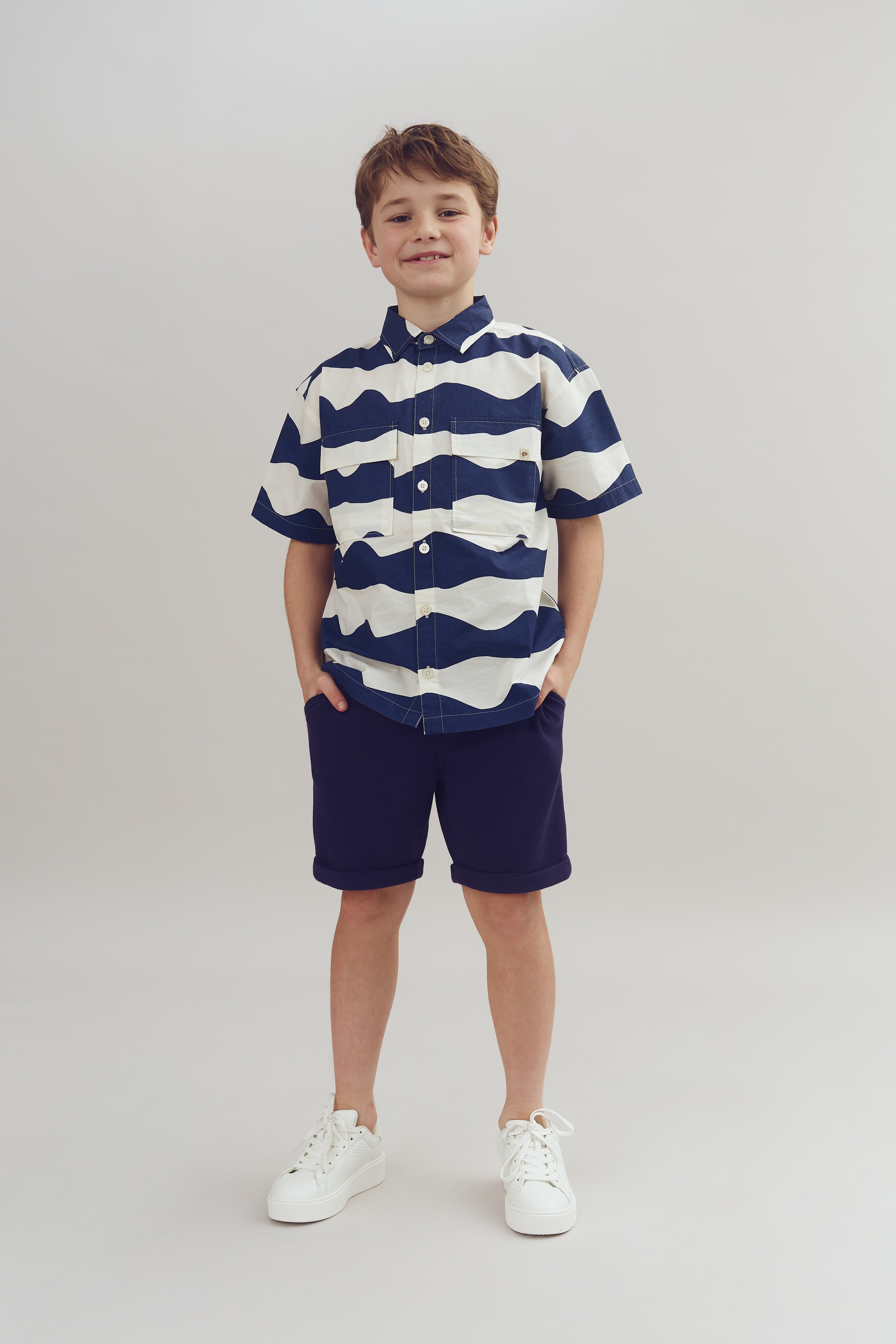 Junge in gestreiftem Hemd und blauen Shorts steht mit Händen in den Taschen, weißem Hintergrund. Trägt weiße Sneakers, lächelt leicht.