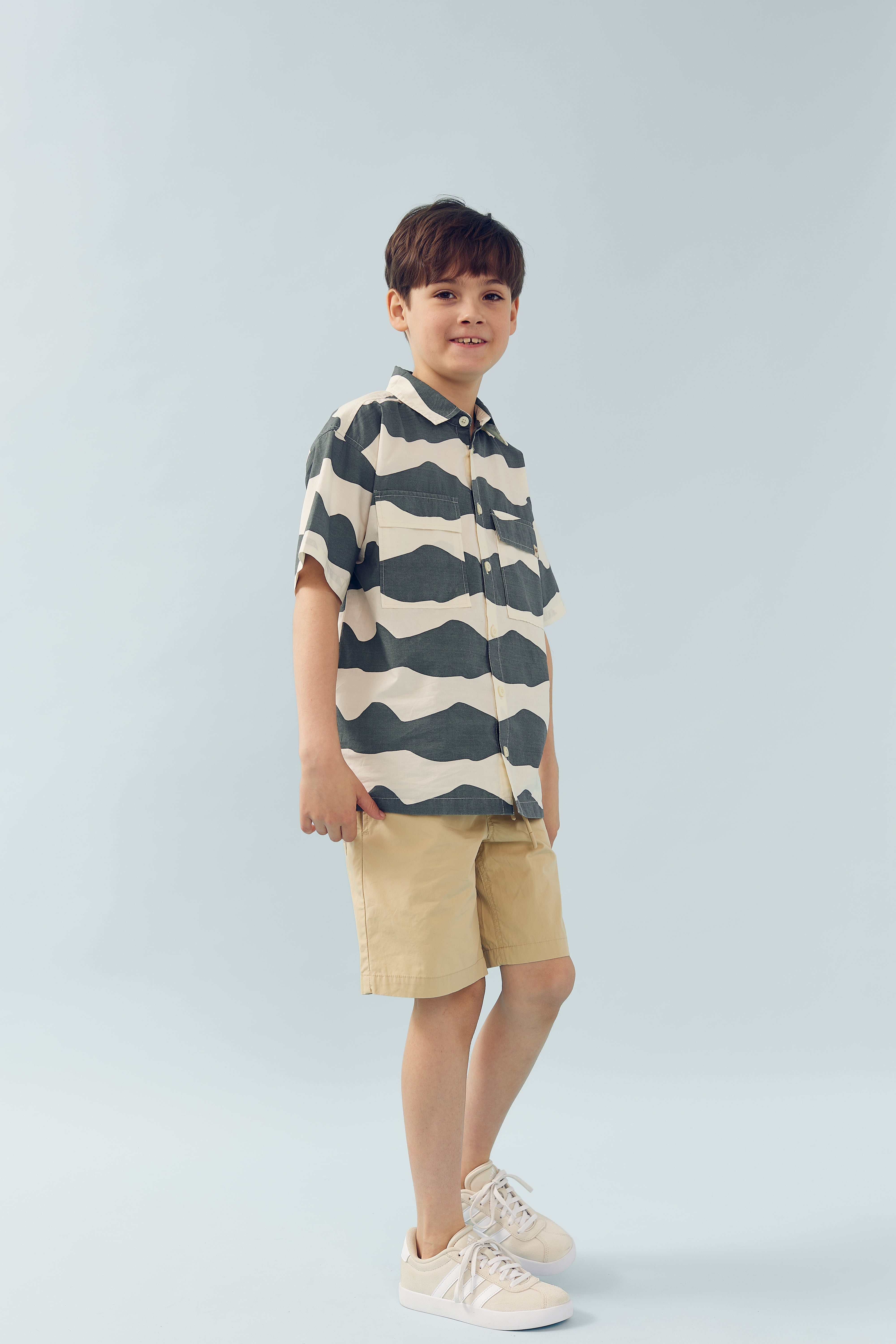 Ein Junge steht lächelnd, trägt ein gestreiftes Hemd, beige Shorts und weiße Turnschuhe vor einem hellblauen Hintergrund.