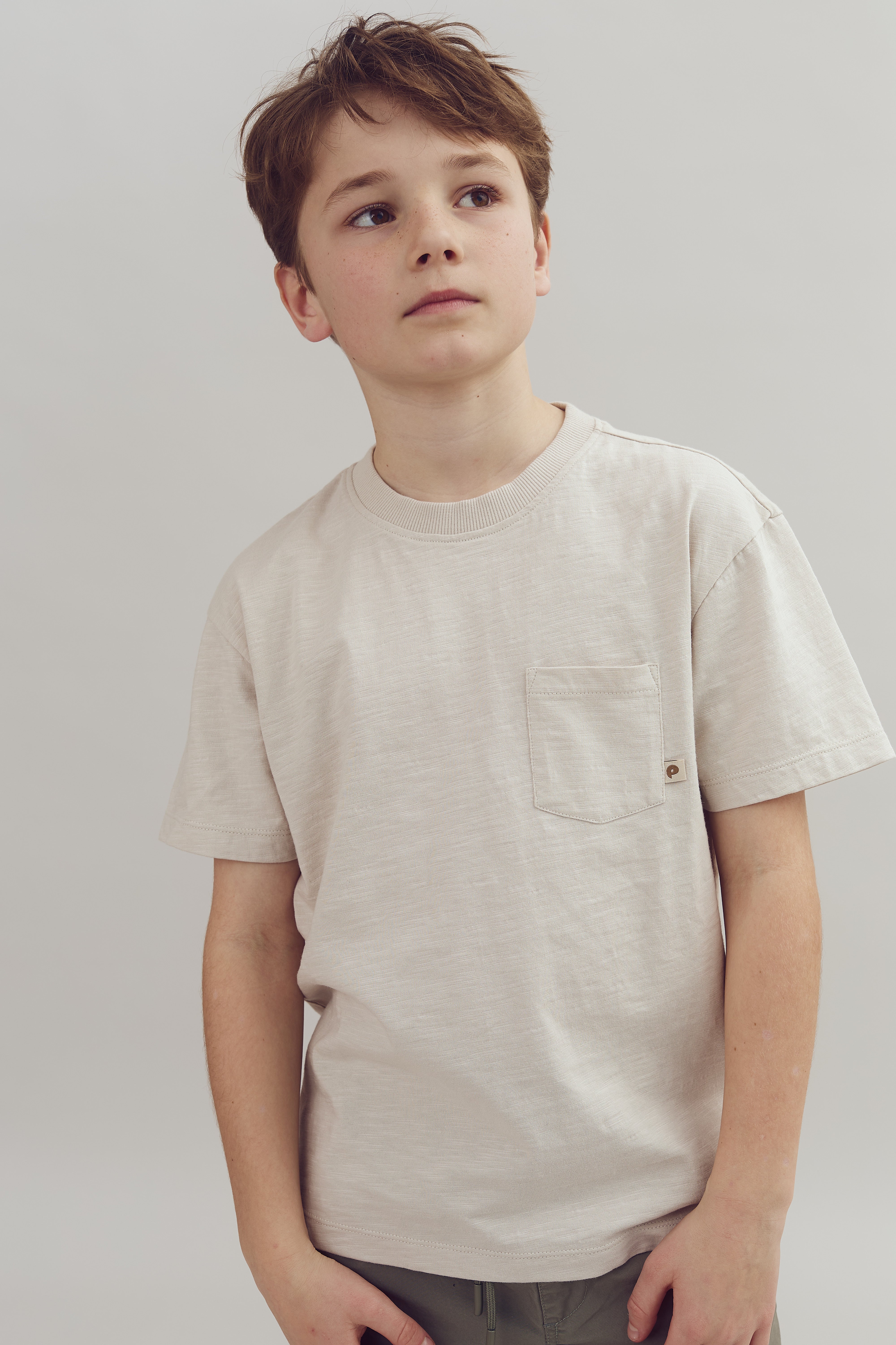 Ein Junge schaut nachdenklich zur Seite, trägt ein beiges T-Shirt mit Brusttasche, in einem neutralen Hintergrund.