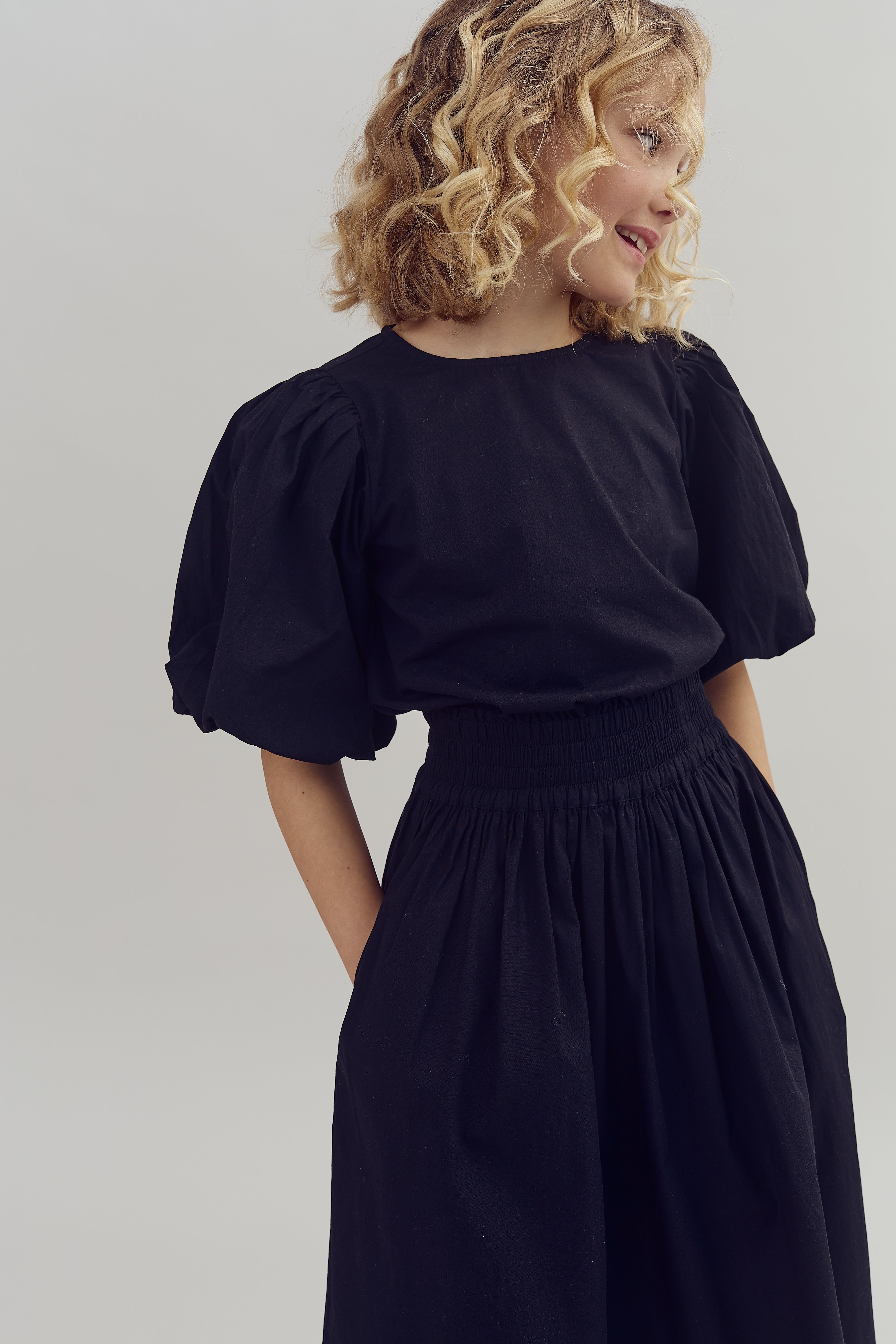 Ein Kind mit lockigem blondem Haar trägt ein schwarzes Kleid mit Puffärmeln und blickt zur Seite, in einem neutralen, hellen Hintergrund.