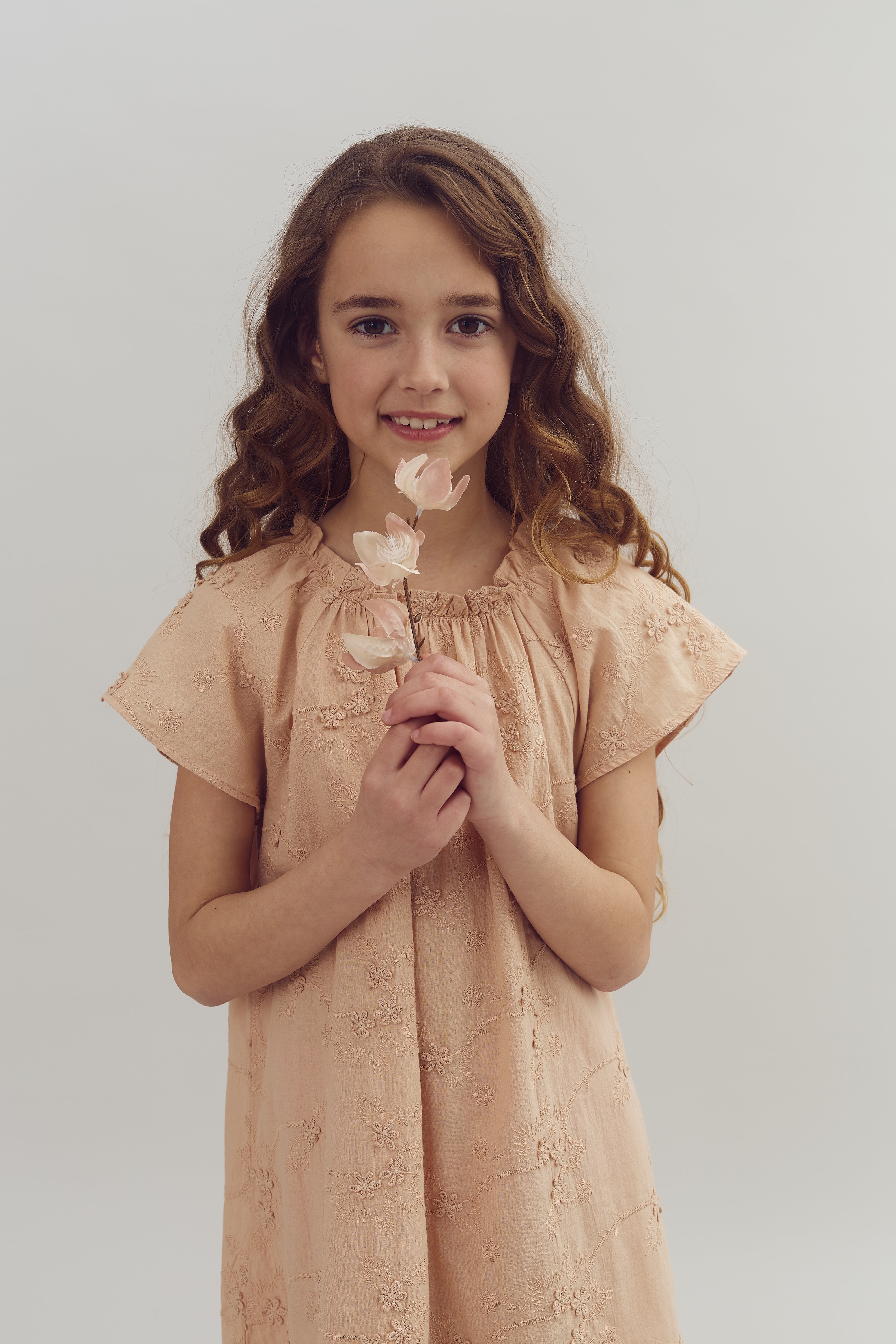 Mädchen hält zartrosa Blume in Händen, lächelt sanft. Sie trägt ein pastellrosa Kleid mit Blumenmuster. Der Hintergrund ist neutral, wodurch das Kind im Mittelpunkt steht.