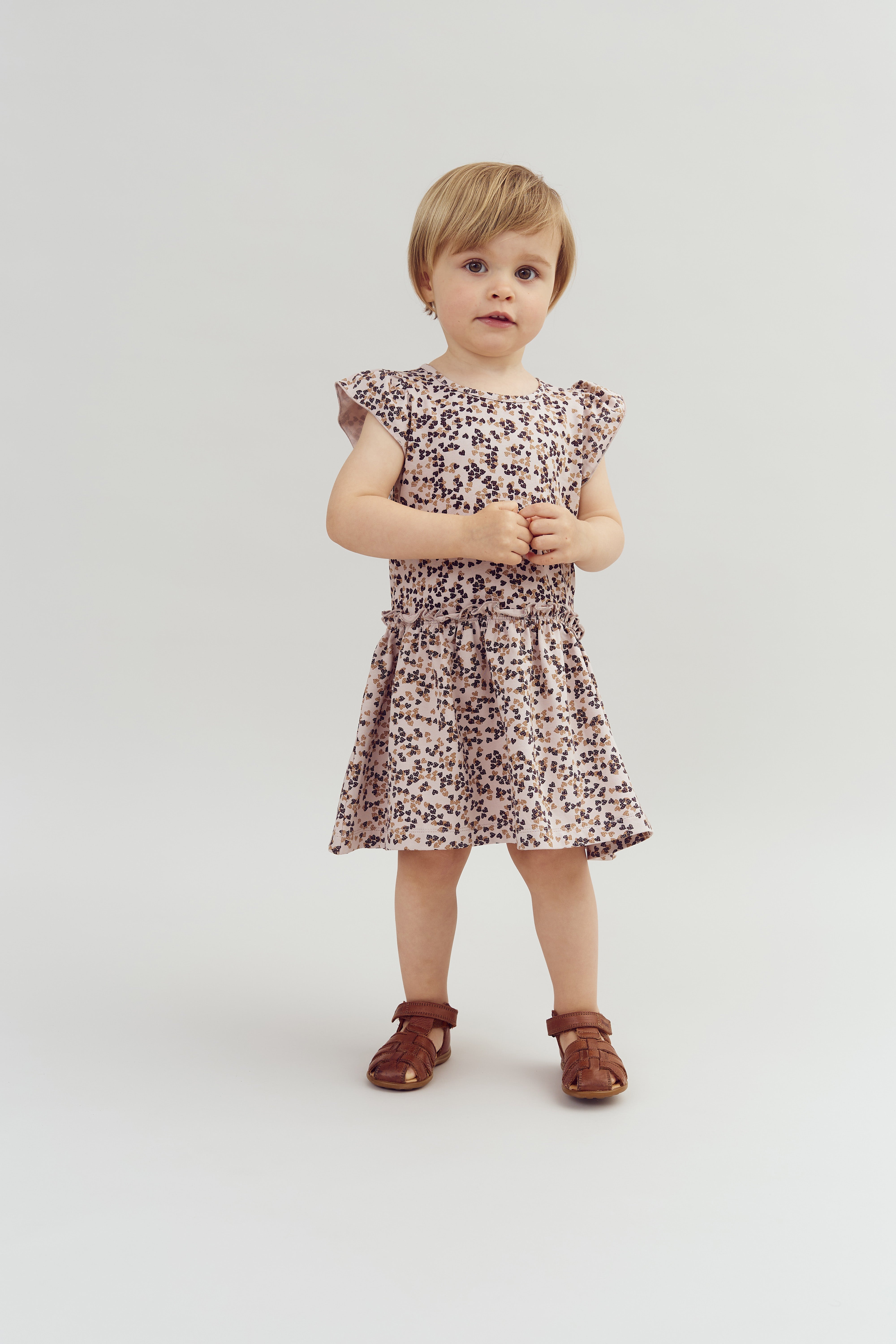 Ein Kleinkind steht in einem Kleid mit Blumenmuster und braunen Sandalen auf einem hellen Hintergrund und schaut ruhig in die Kamera.