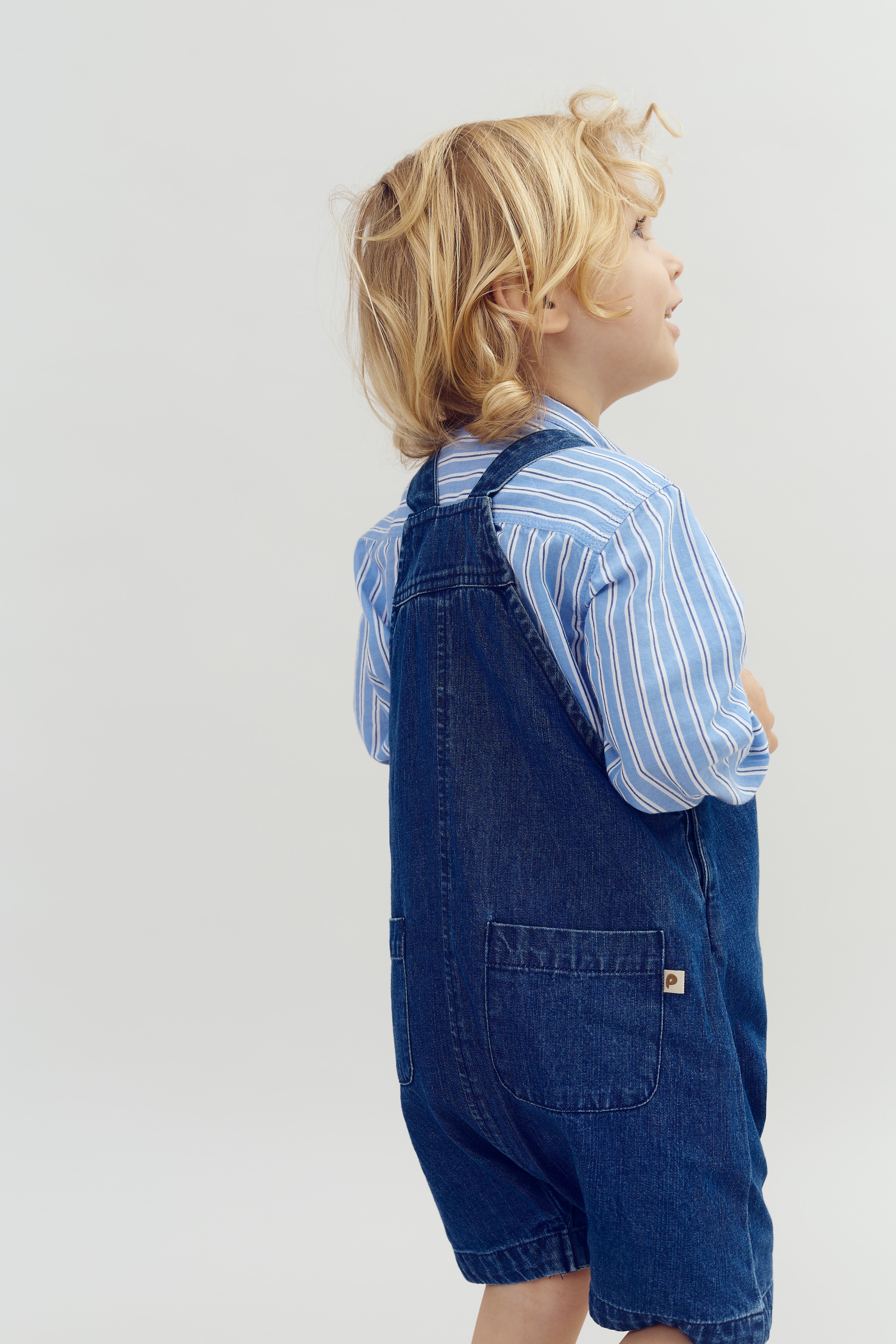 Ein Kind steht seitlich, trägt einen blau-weiß gestreiften Pullover und eine blaue Jeans-Latzhose, vor einem neutralen, hellen Hintergrund.