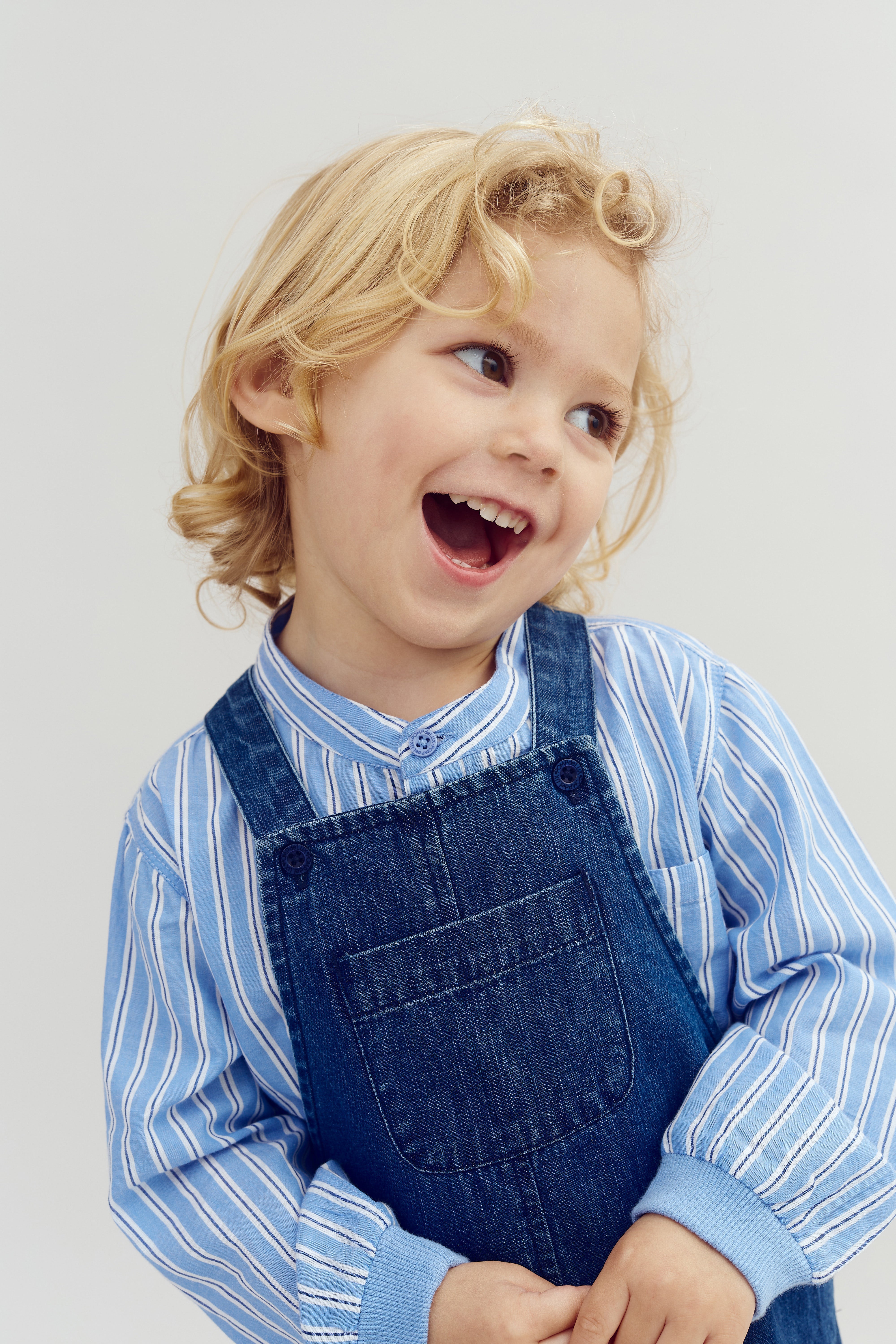 Ein lachendes Kind mit lockigem blondem Haar trägt einen blau gestreiften Pullover und eine Jeans-Latzhose. Der Hintergrund ist schlicht und einfarbig.