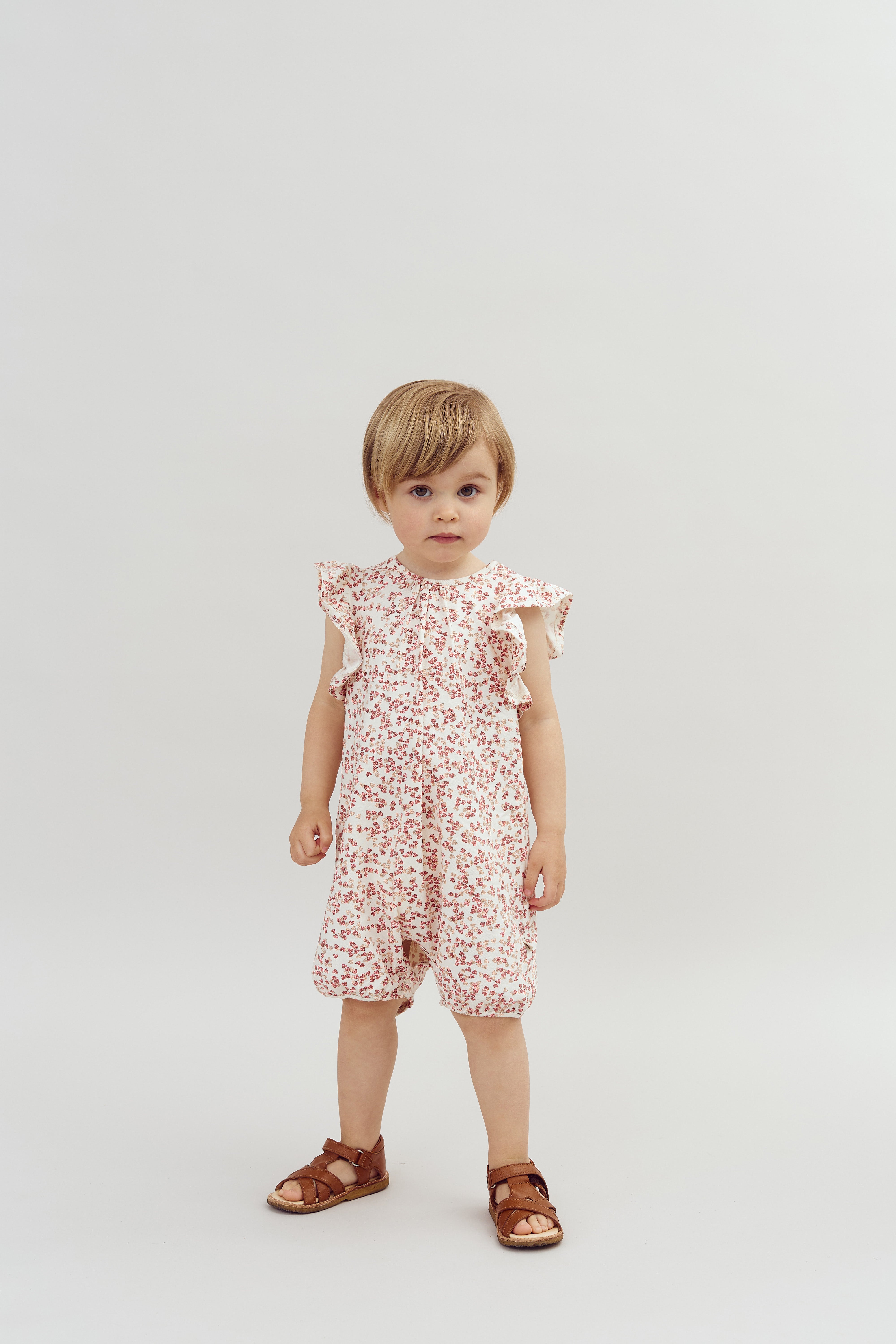 Ein Kind steht in einem gemusterten Sommerkleid mit Flügelärmeln und braunen Sandalen vor einem neutralen, hellgrauen Hintergrund.