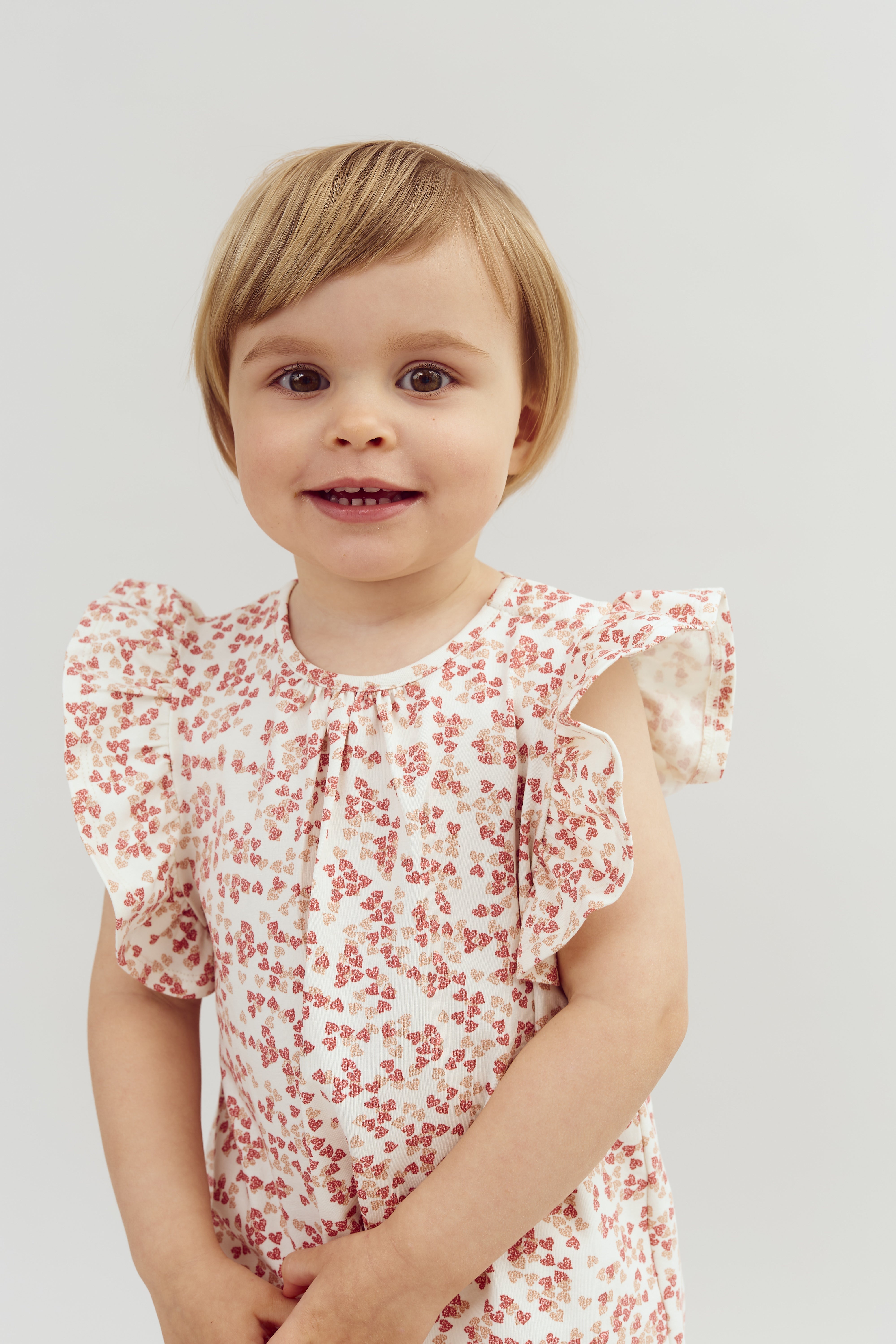 Kleinkind lächelt ruhig, trägt ein weißes Kleid mit roten Blumenmustern, steht vor einem neutralen grauen Hintergrund.