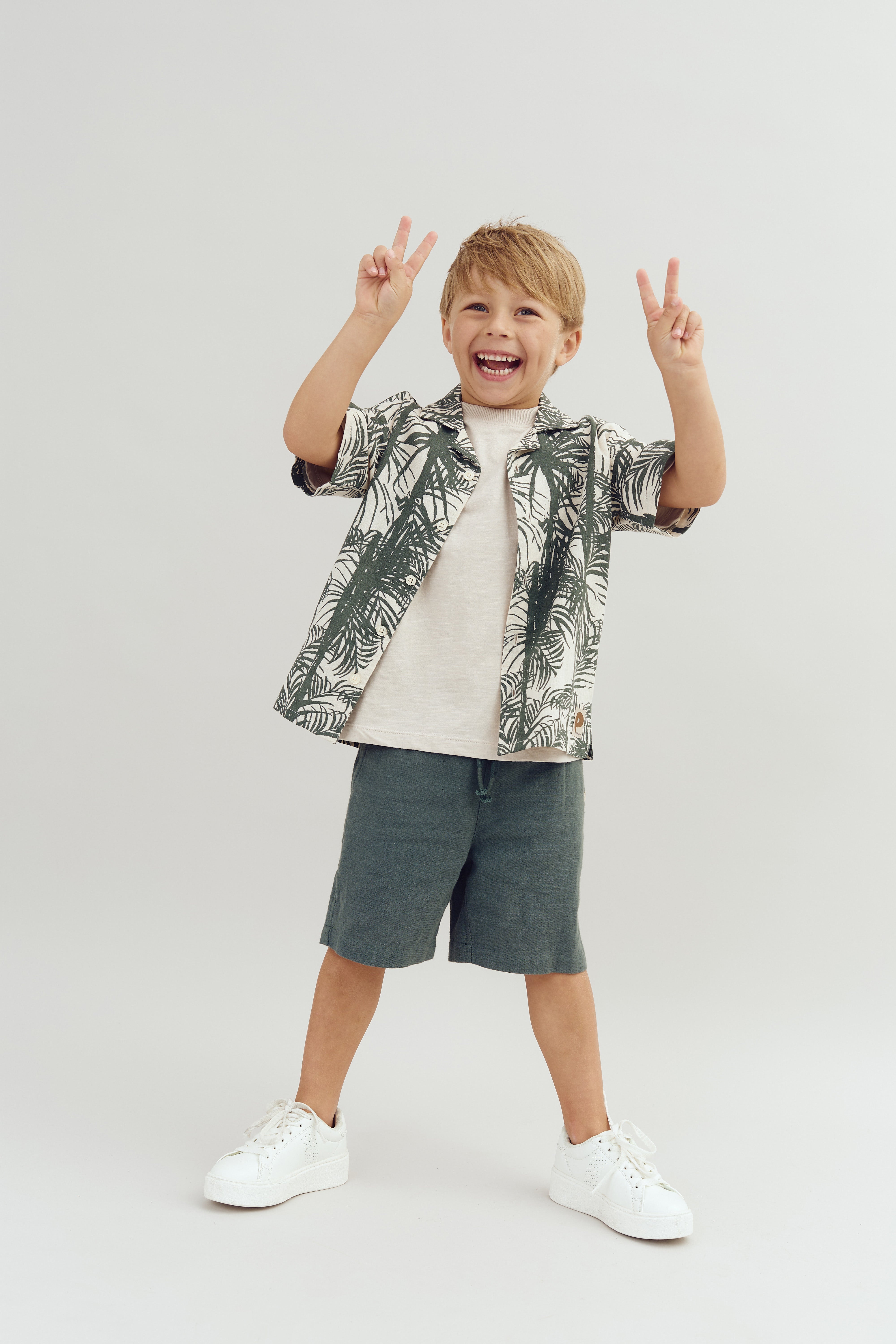 Ein Junge lächelt und zeigt mit beiden Händen das Peace-Zeichen. Er trägt ein Hemd mit Blattmuster, ein helles T-Shirt und dunkle Shorts vor weißem Hintergrund.