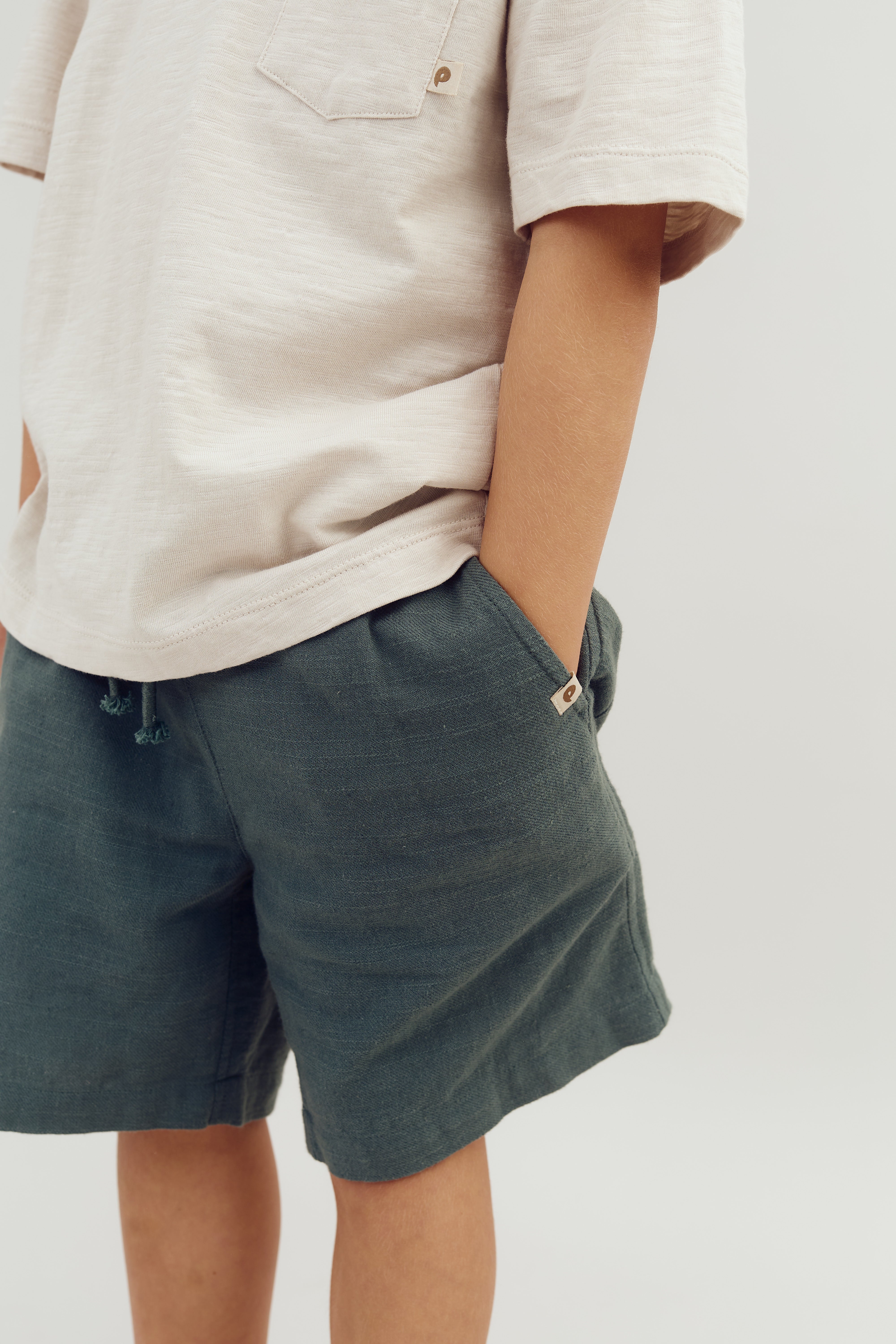 Ein Kind trägt ein beigefarbenes T-Shirt mit Brusttasche und dunkle Shorts, in die Hände in den Taschen steckend. Der Hintergrund ist schlicht und minimalistisch.
