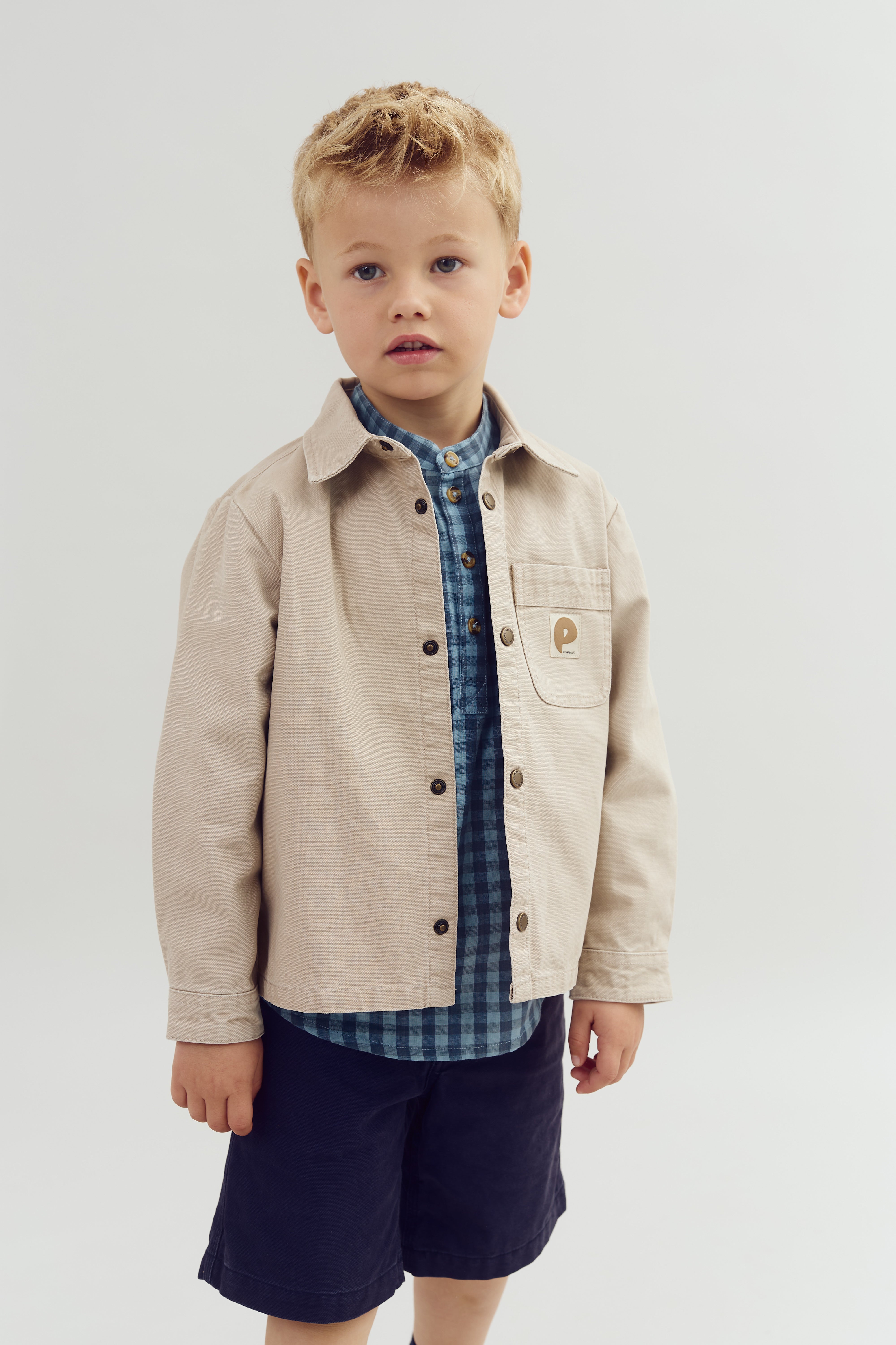 Ein Junge steht entspannt, trägt eine beige Jacke mit einem Logo "P", ein blau-kariertes Hemd und dunkelblaue Shorts vor neutralem, hellem Hintergrund.
