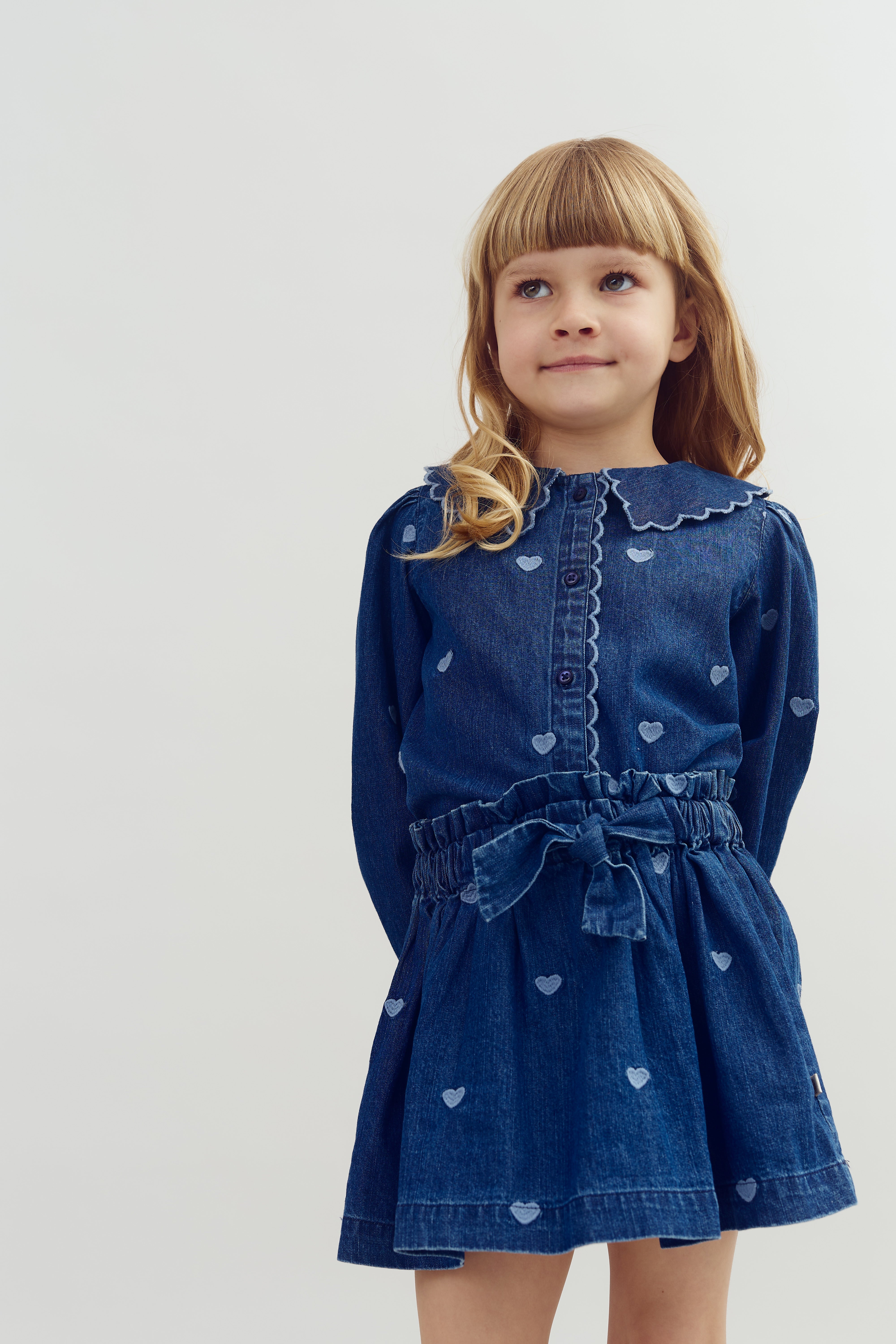 Mädchen trägt ein blaues Jeanskleid mit Herzmustern, steht still und lächelt leicht in einem hellen, neutralen Raum.
