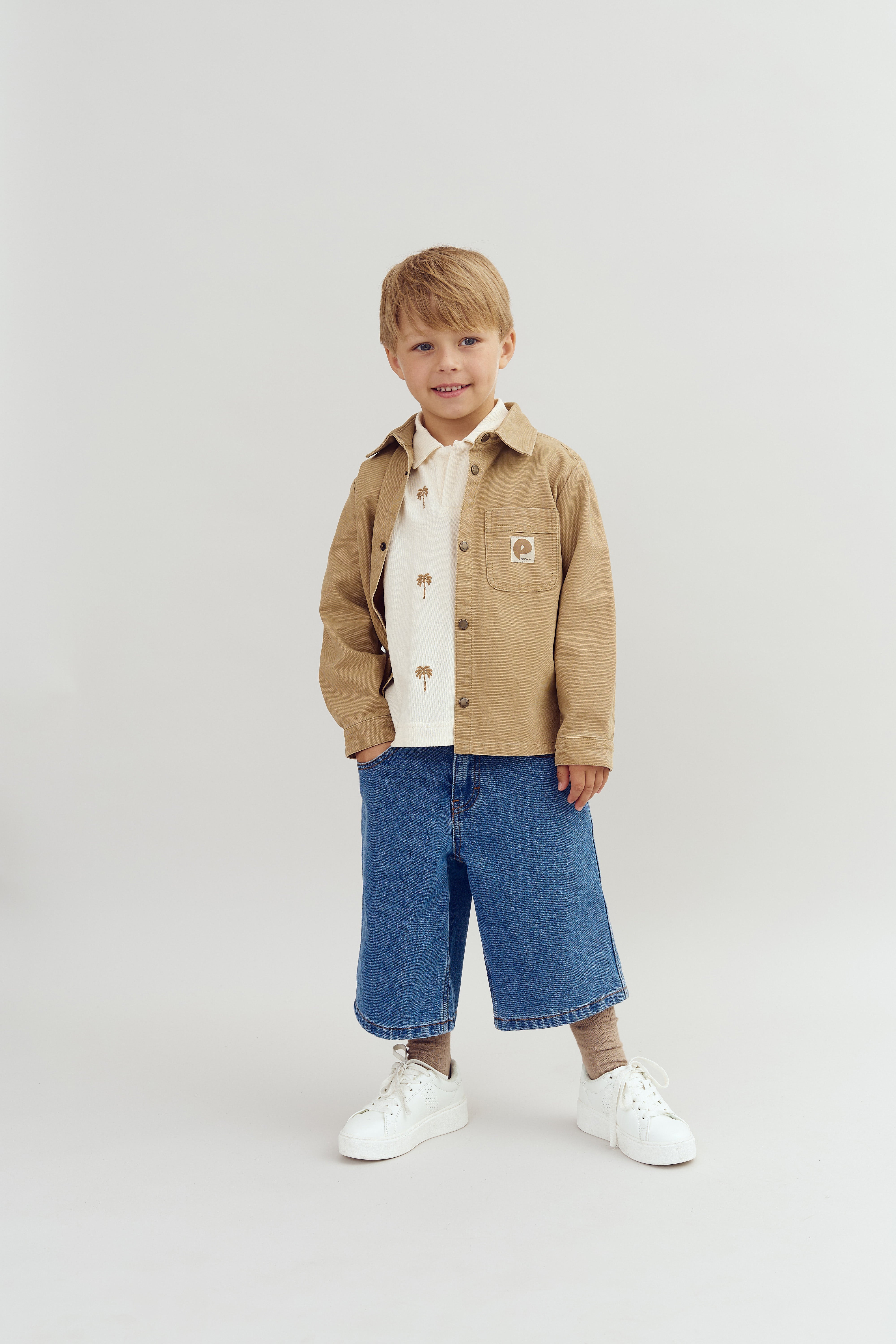 Ein Junge steht lächelnd im Studio, trägt eine beige Jacke, ein bedrucktes Hemd und weite, blaue Jeans. Der Hintergrund ist schlicht und hell.