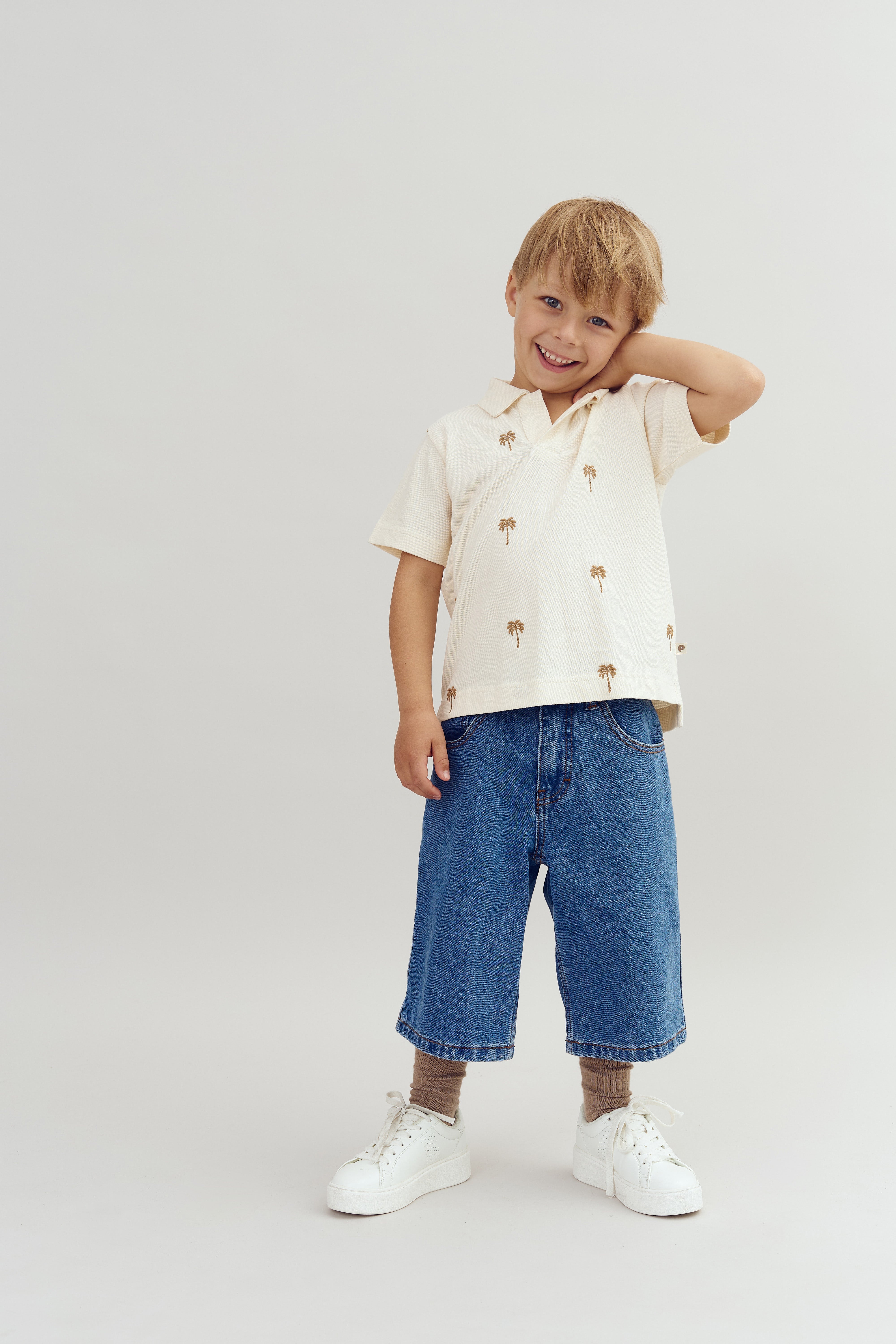 Ein Junge steht lächelnd in einem hellen Raum. Er trägt ein beiges Hemd mit Palmenmotiven, weite blaue Jeans, braune Socken und weiße Turnschuhe.