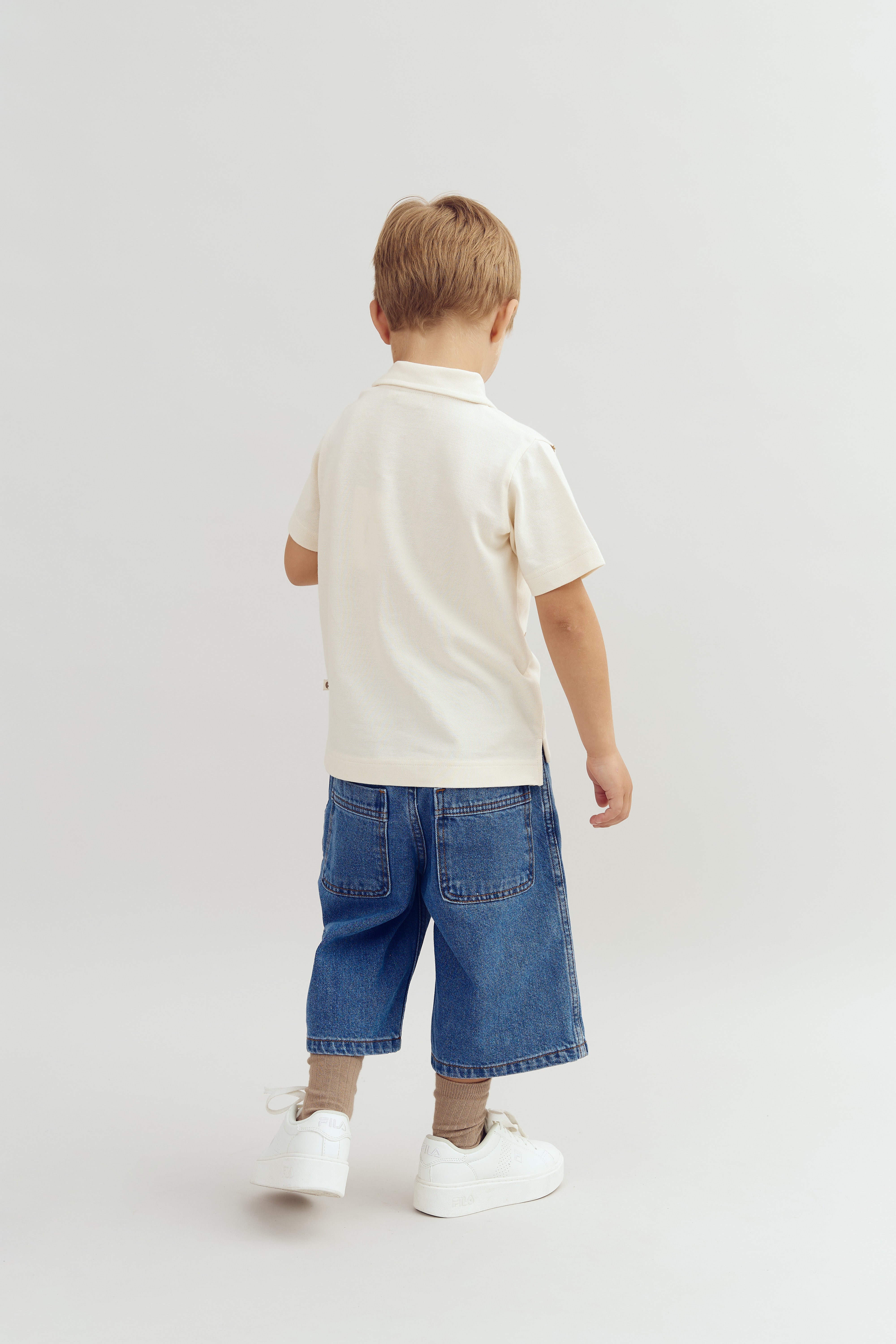 Ein Kind steht, den Rücken zur Kamera, in einem weißen T-Shirt, blauen Jeans und weißen Turnschuhen, gegen einen neutralen, hellen Hintergrund.