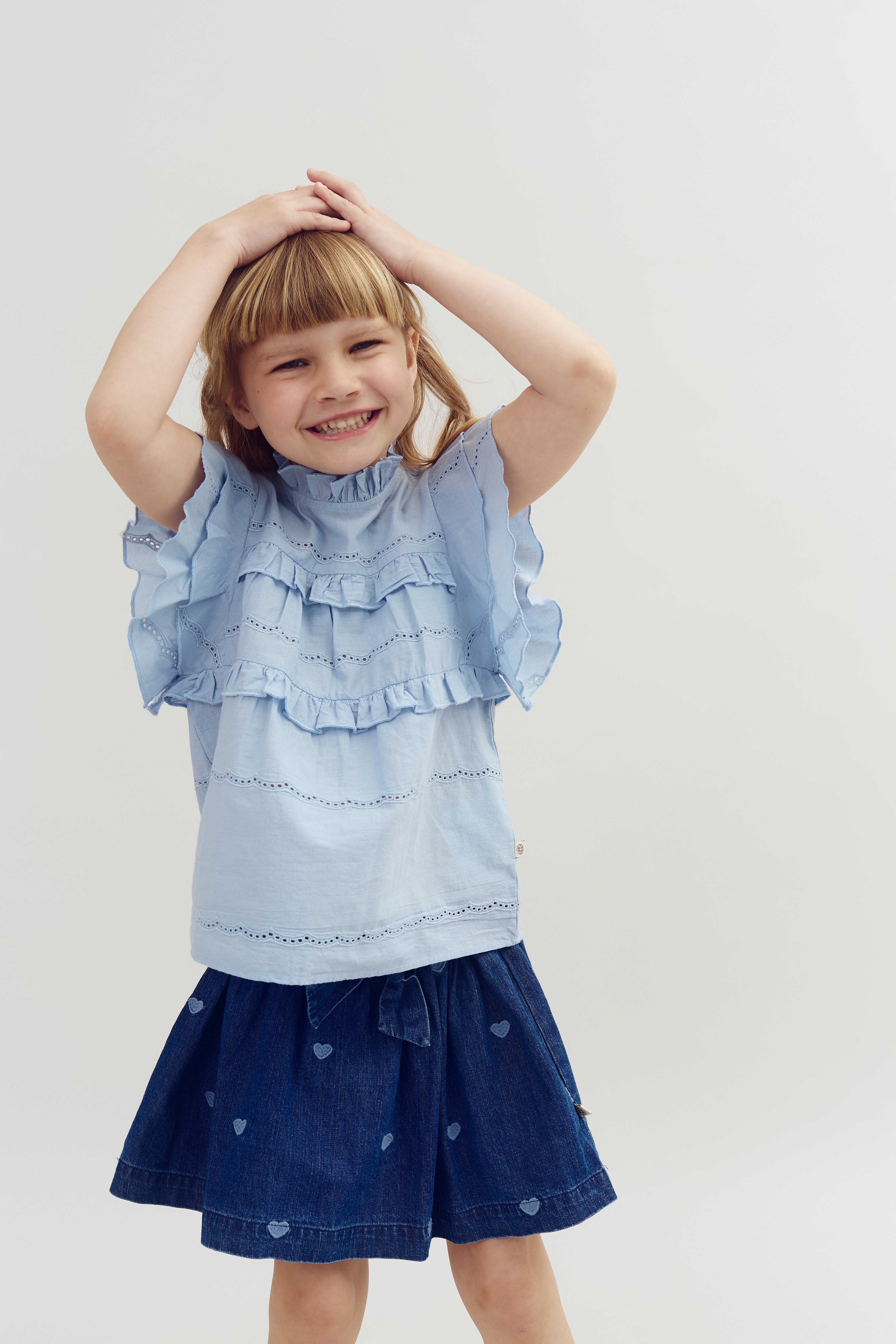 Ein Kind lächelt, hält die Hände auf dem Kopf und trägt ein hellblaues Shirt mit Rüschen und einen Jeansrock mit Herzmotiven. Der Hintergrund ist schlicht und weiß.