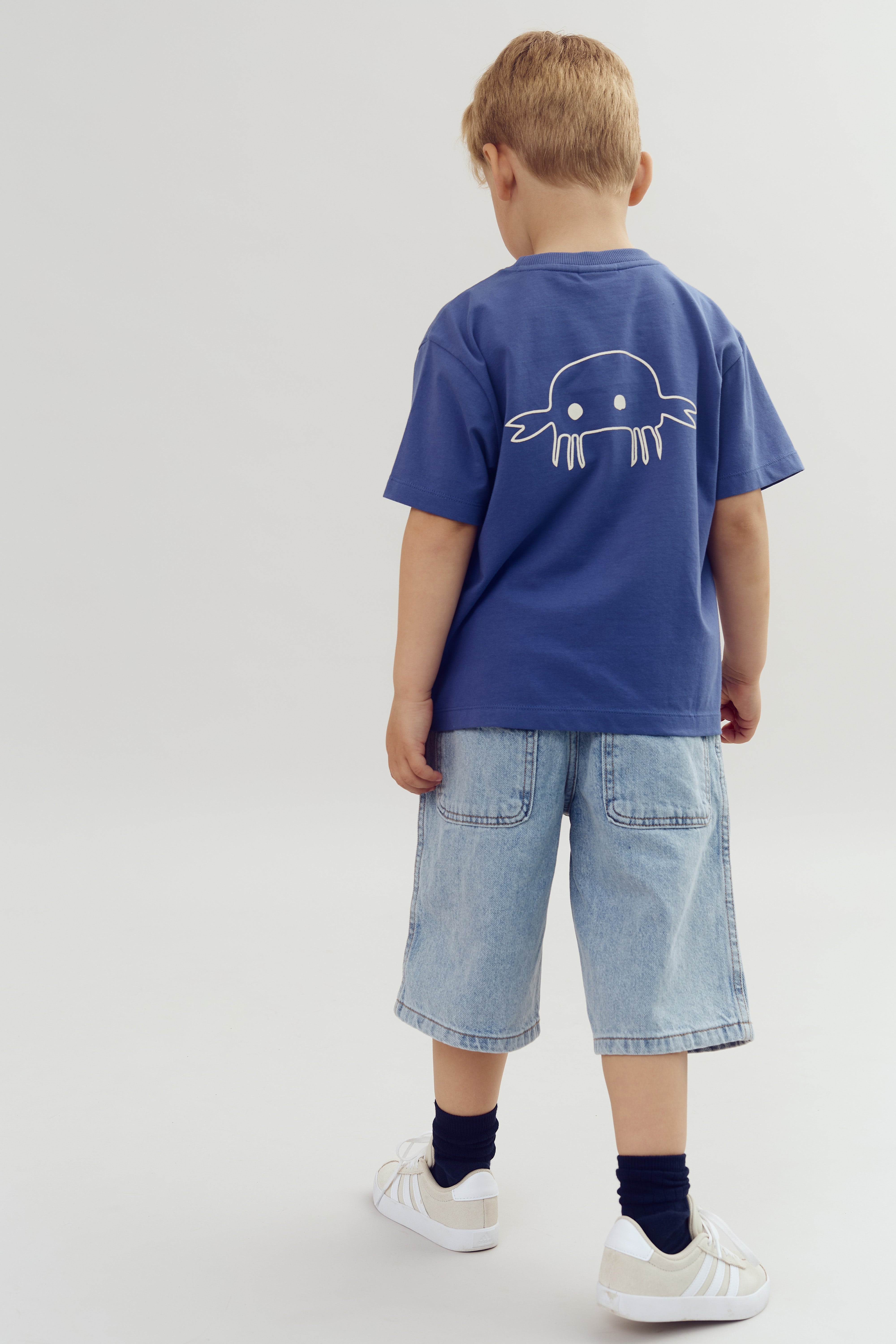 Ein Kind trägt ein blaues T-Shirt mit einem Krabbenmotiv und Jeansshorts. Es steht in einem schlichten, hellen Raum. Weiße Sneakers und dunkle Socken vervollständigen das Outfit.