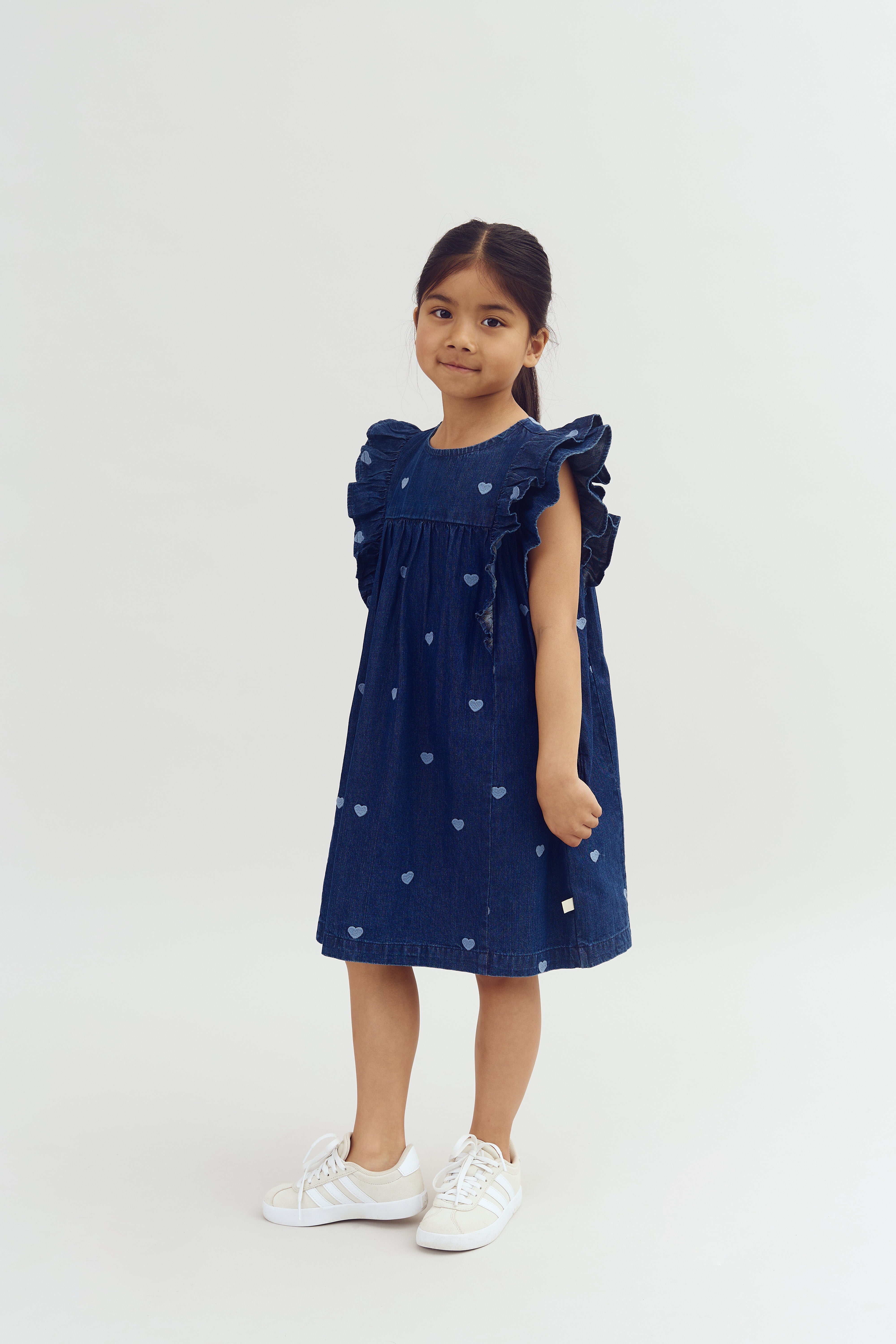 Ein Mädchen trägt ein blaues Kleid mit herzförmigen Mustern, steht ruhig und schaut in die Kamera. Sie trägt weiße Turnschuhe. Der Hintergrund ist schlicht und hellgrau.