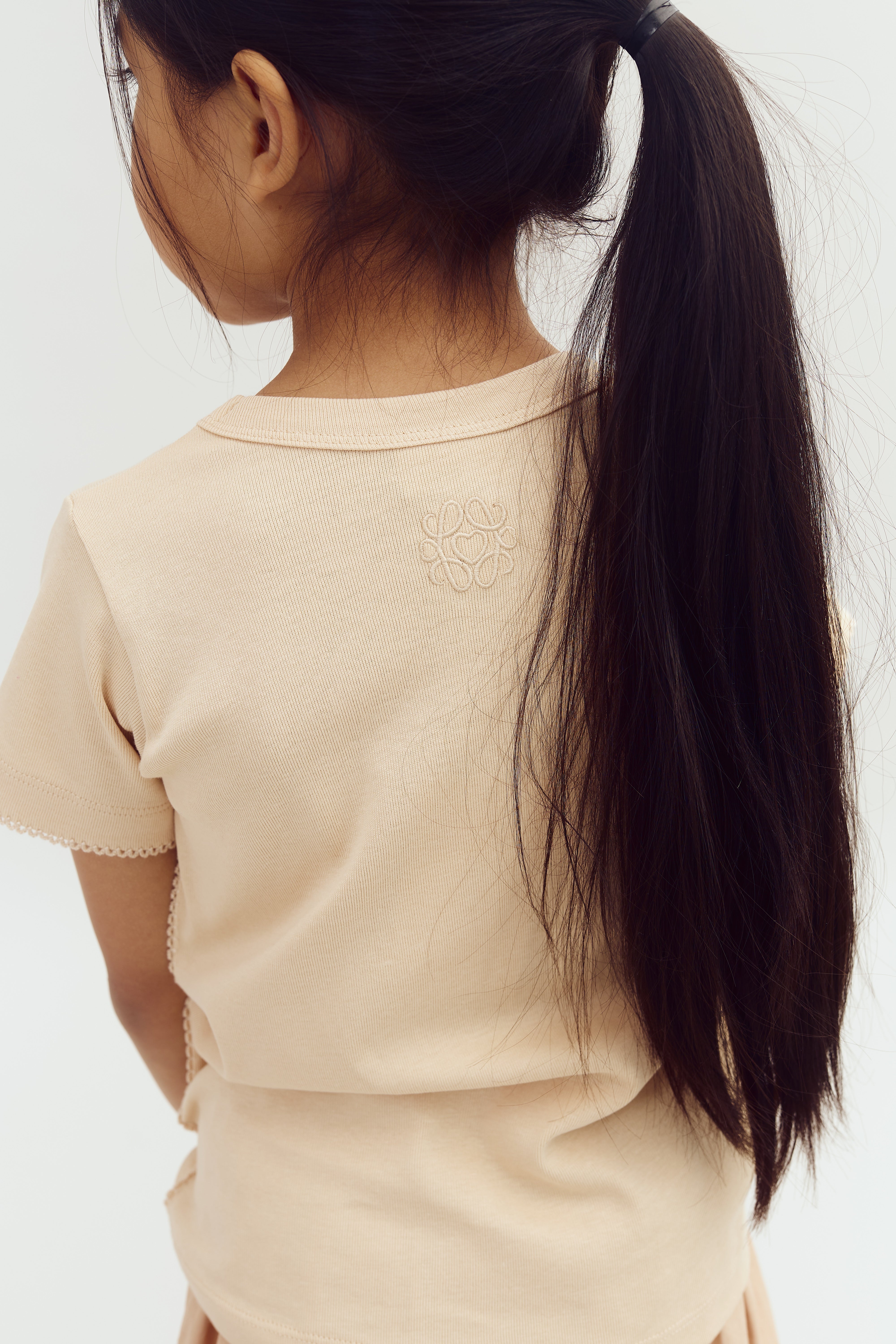 Ein Mädchen mit langen, dunklen Haaren im Pferdeschwanz steht seitlich. Sie trägt ein beiges T-Shirt mit einem dezenten Muster am Rücken, vor neutralem Hintergrund.