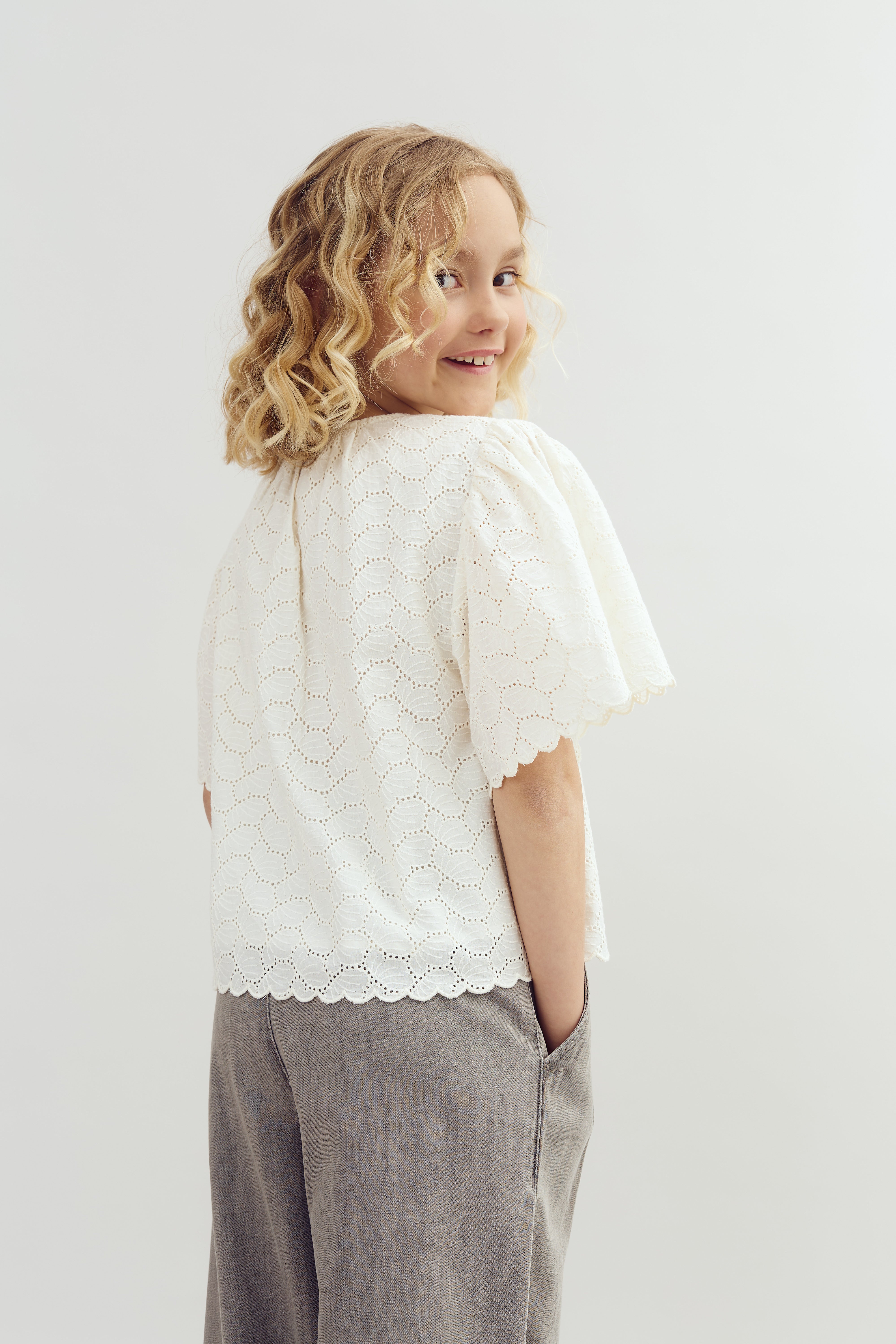 Ein Mädchen mit lockigem blondem Haar lächelt, trägt eine weiße Spitze-Bluse und graue Hosen, mit der Hand in der Tasche; der Hintergrund ist schlicht und hell.
