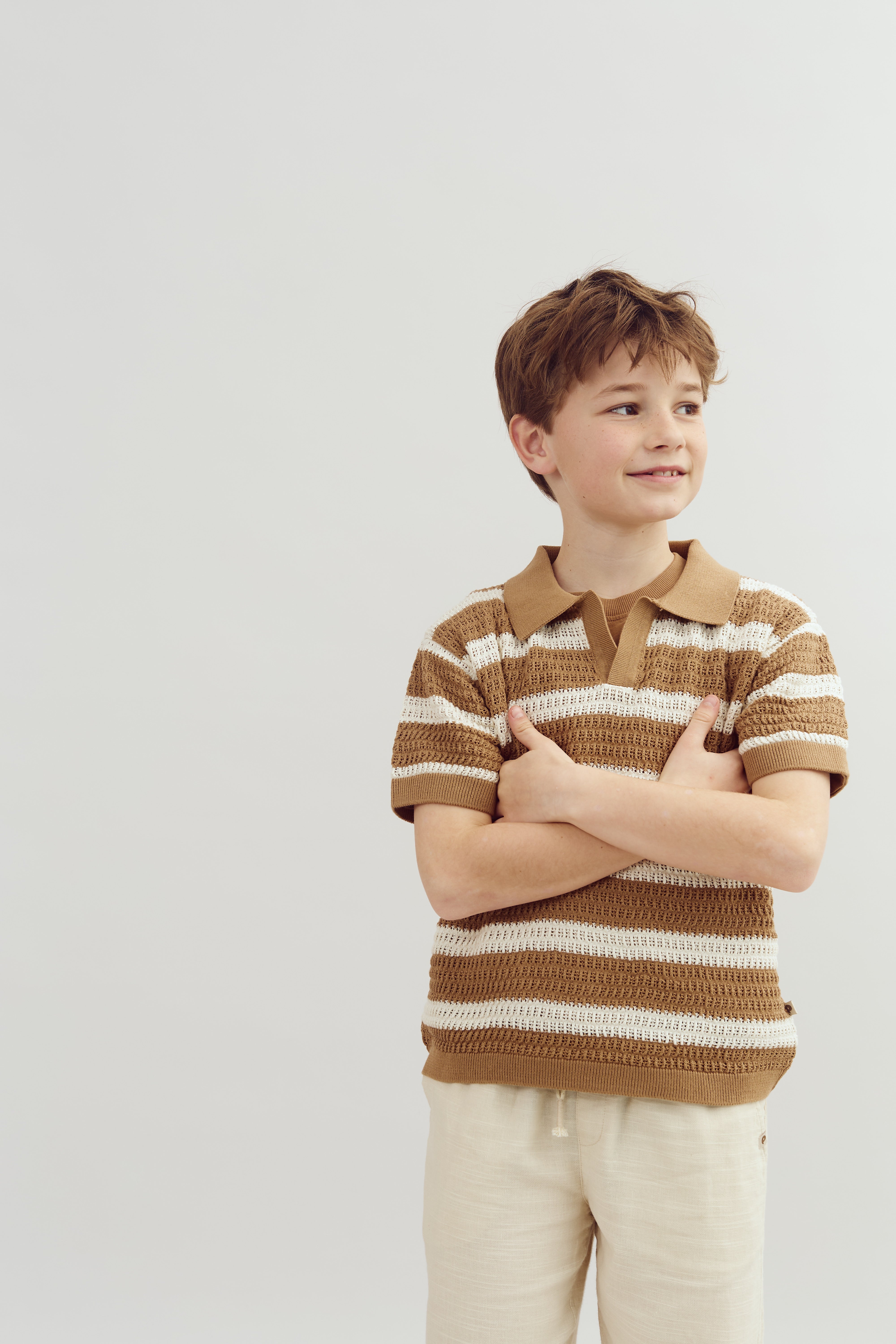 Ein Junge steht lächelnd mit verschränkten Armen. Er trägt ein gestreiftes, braun-weißes Poloshirt und helle Hosen. Der Hintergrund ist einfarbig und hell.