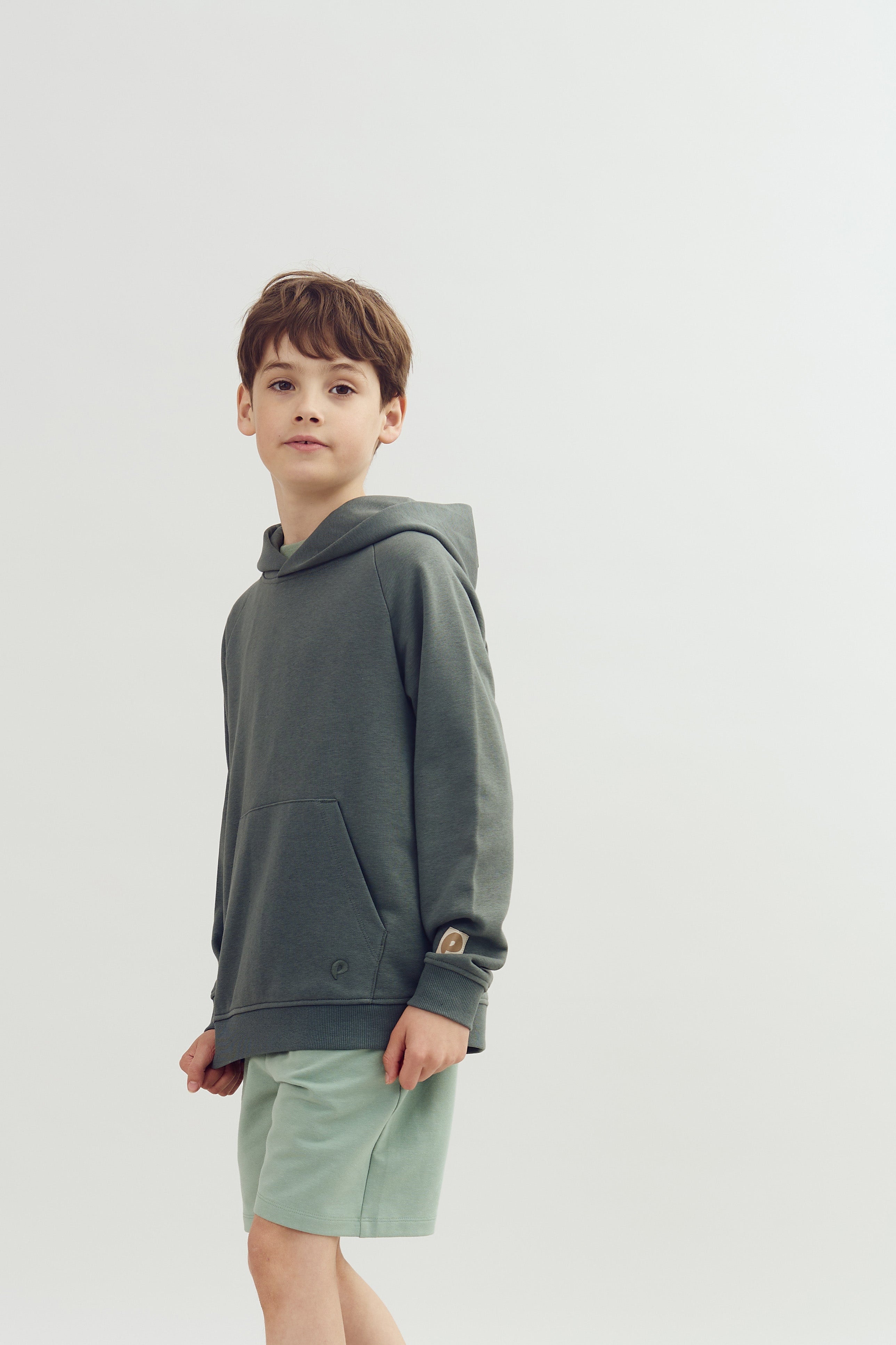 Junge in grauem Kapuzenpullover und mintfarbenen Shorts steht ruhig; neutraler Hintergrund.