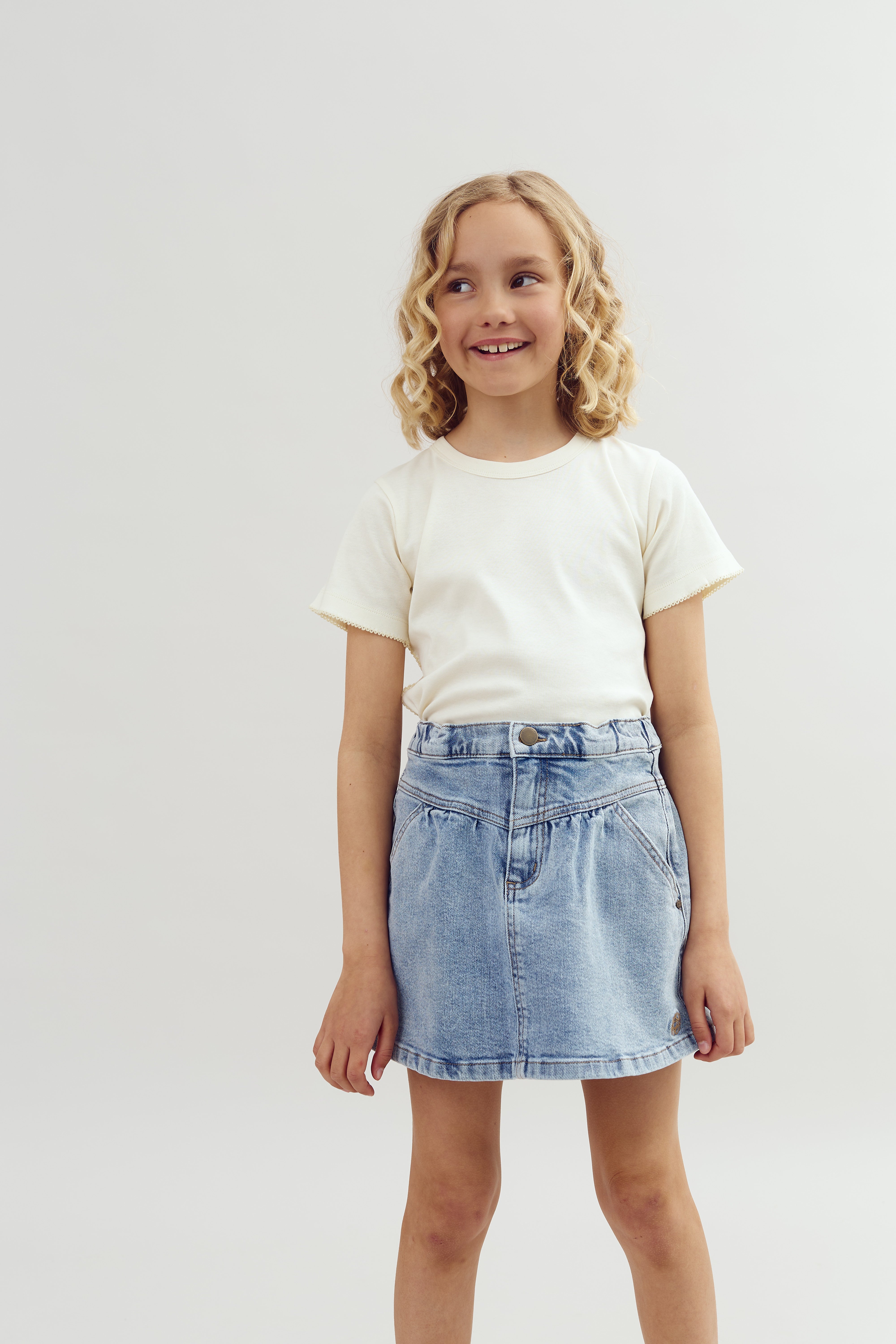 Ein Kind steht lächelnd in einem hellen Raum, trägt ein weißes T-Shirt und einen blauen Jeansrock. Das Kind hat lockiges blondes Haar und schaut zur Seite.