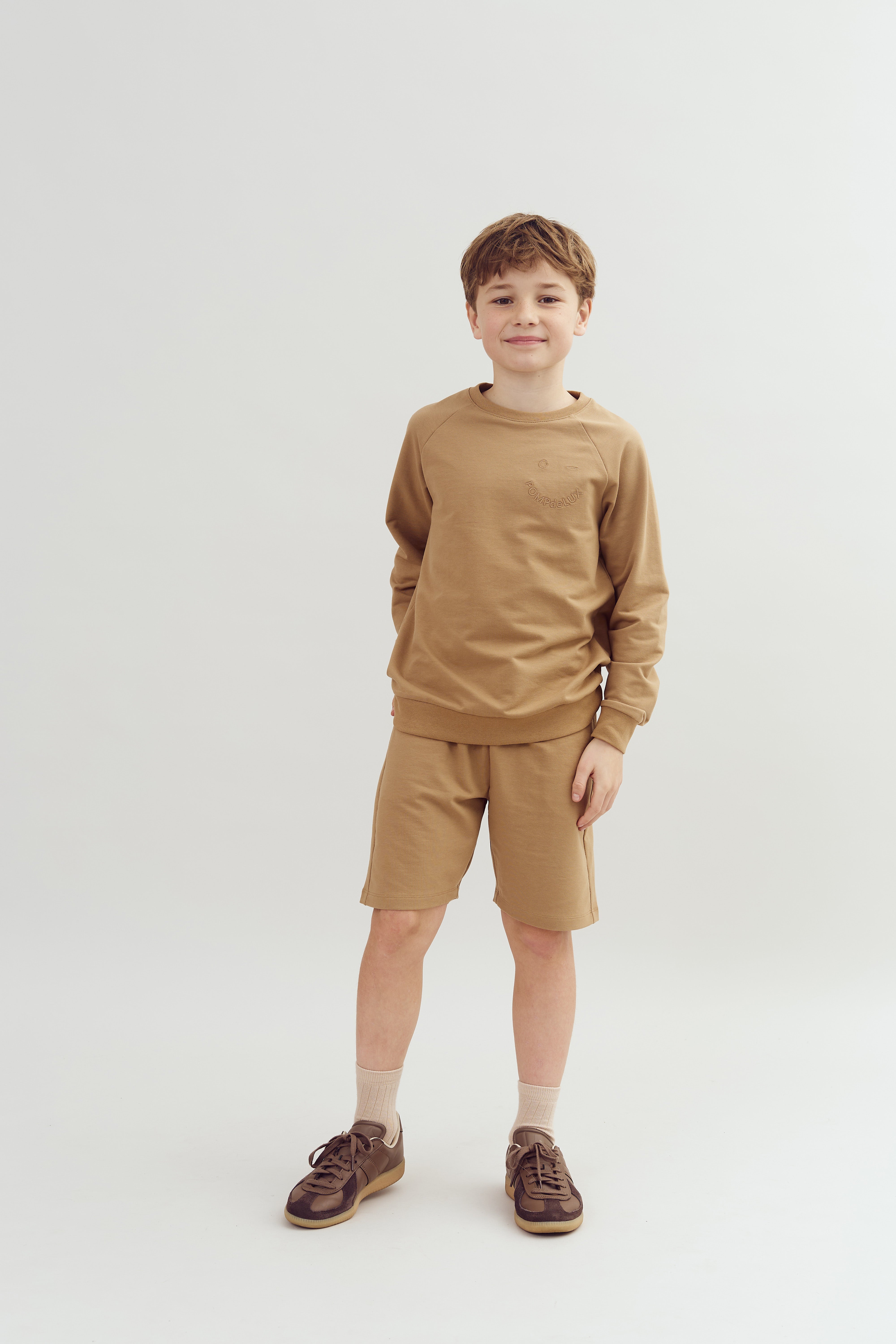 Ein Junge steht lächelnd in einem hellen Raum. Er trägt einen beige-braunen Pullover und passende Shorts sowie braune Turnschuhe auf einem weißen Studiohintergrund.