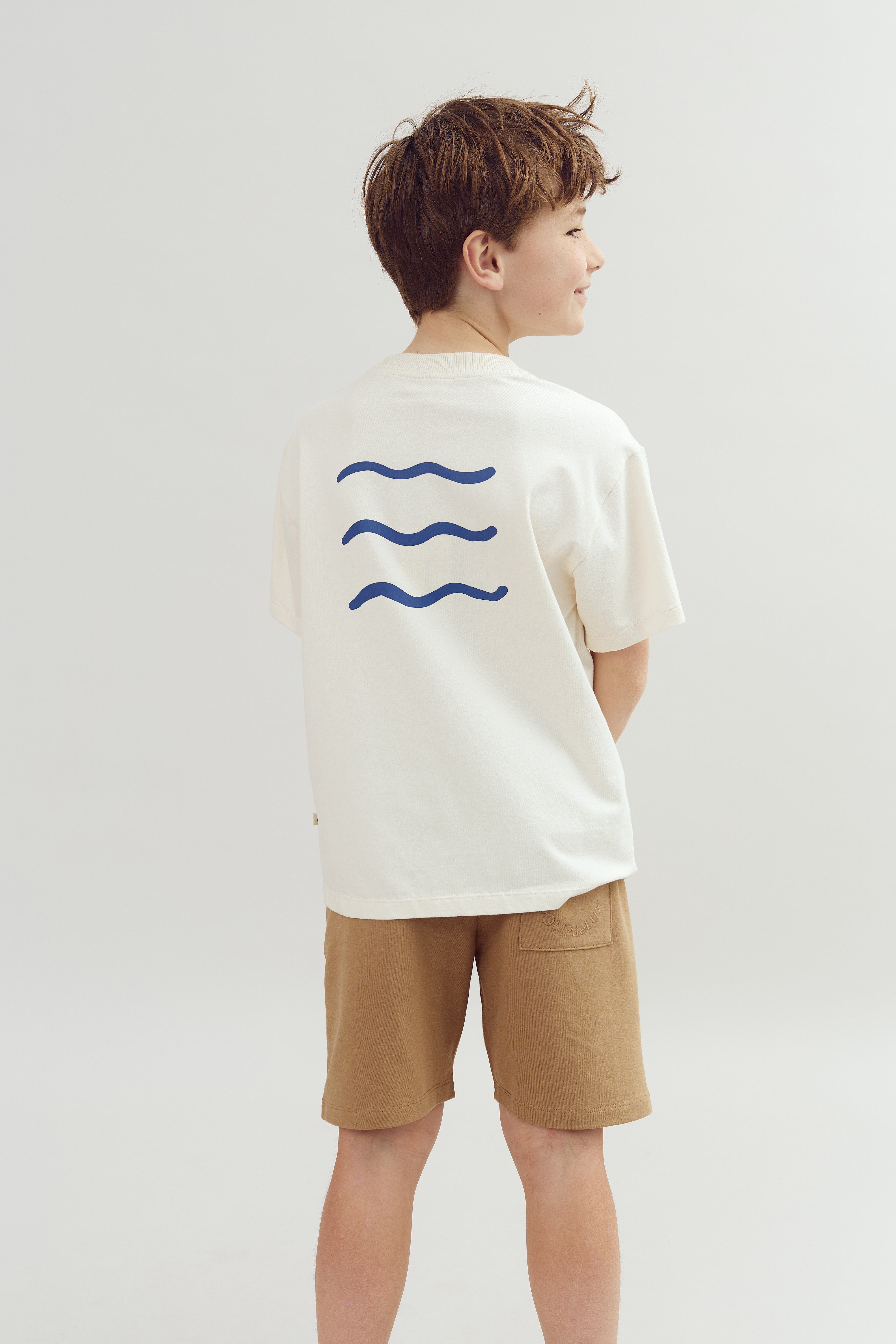 Ein Junge trägt ein weißes T-Shirt mit blauen Wellenlinien auf dem Rücken und braunen Shorts. Er steht seitlich in einem hellen Raum.