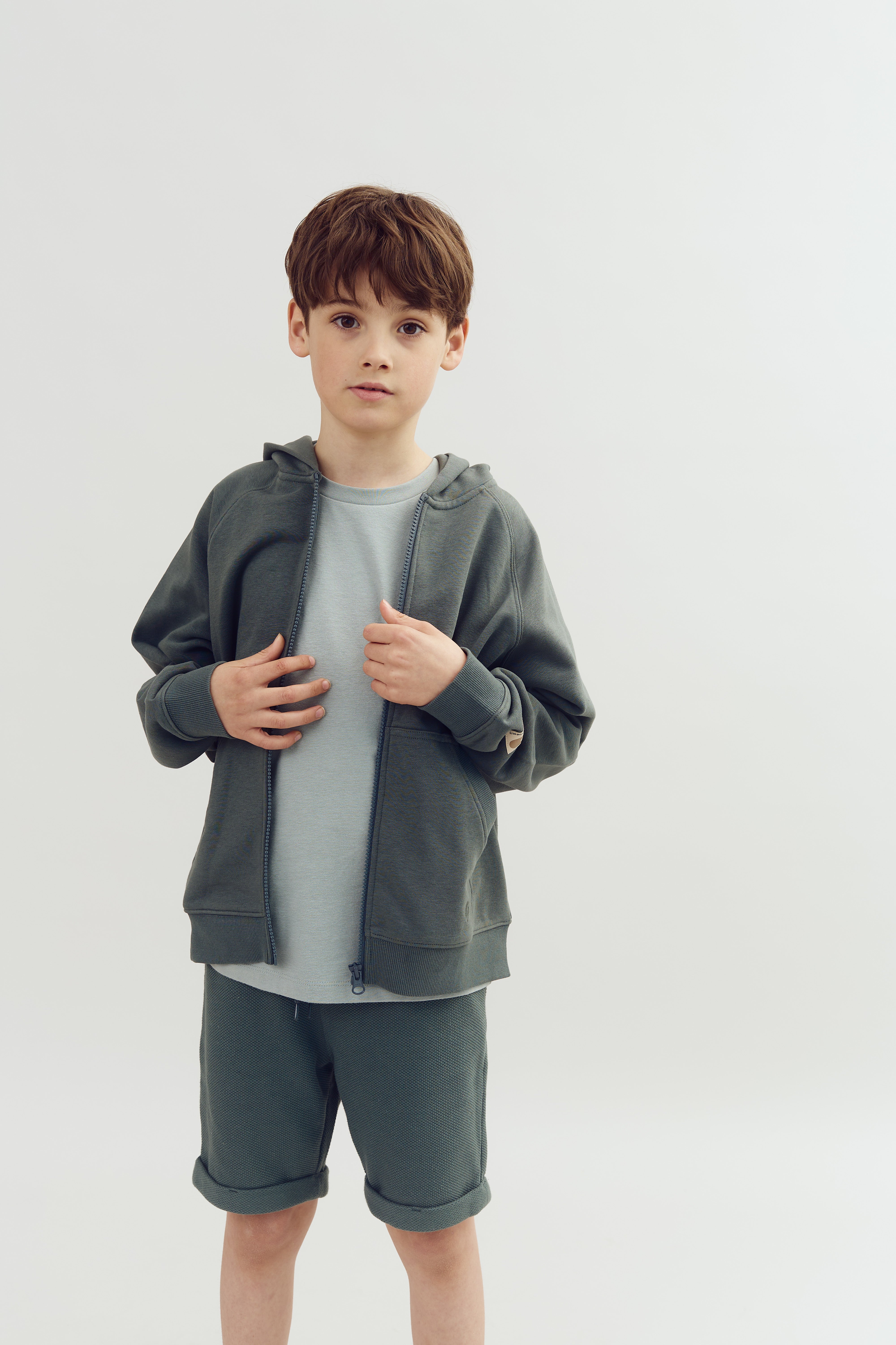 Ein Junge trägt einen grünen Kapuzenpullover und Shorts, hält beide Hände an den Reißverschluss und steht vor einem einfachen, hellen Hintergrund.