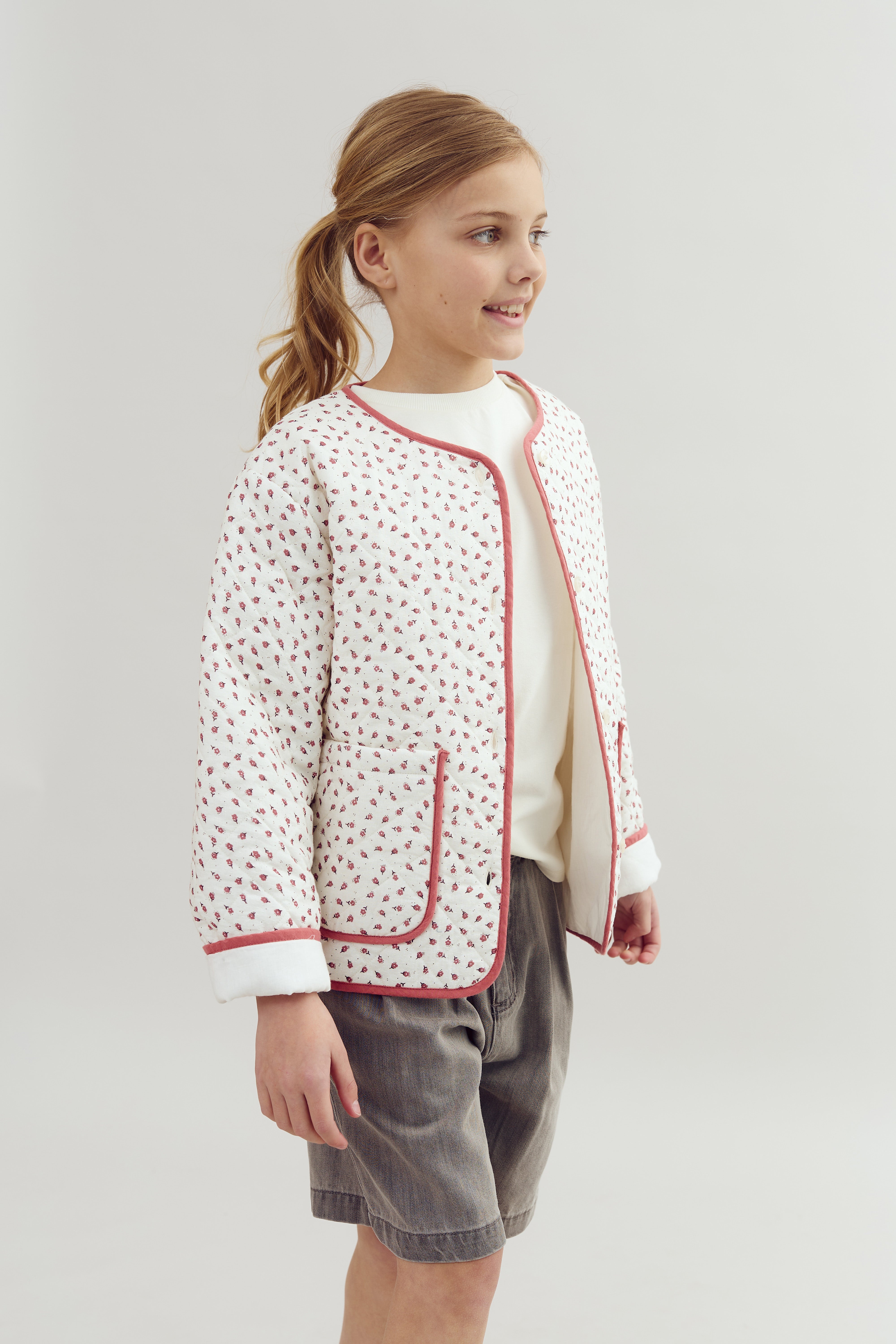 Ein lächelndes Kind trägt eine weiße, gemusterte Jacke mit rosa Kanten und grauen Shorts. Es steht vor einem neutralen, hellen Hintergrund.