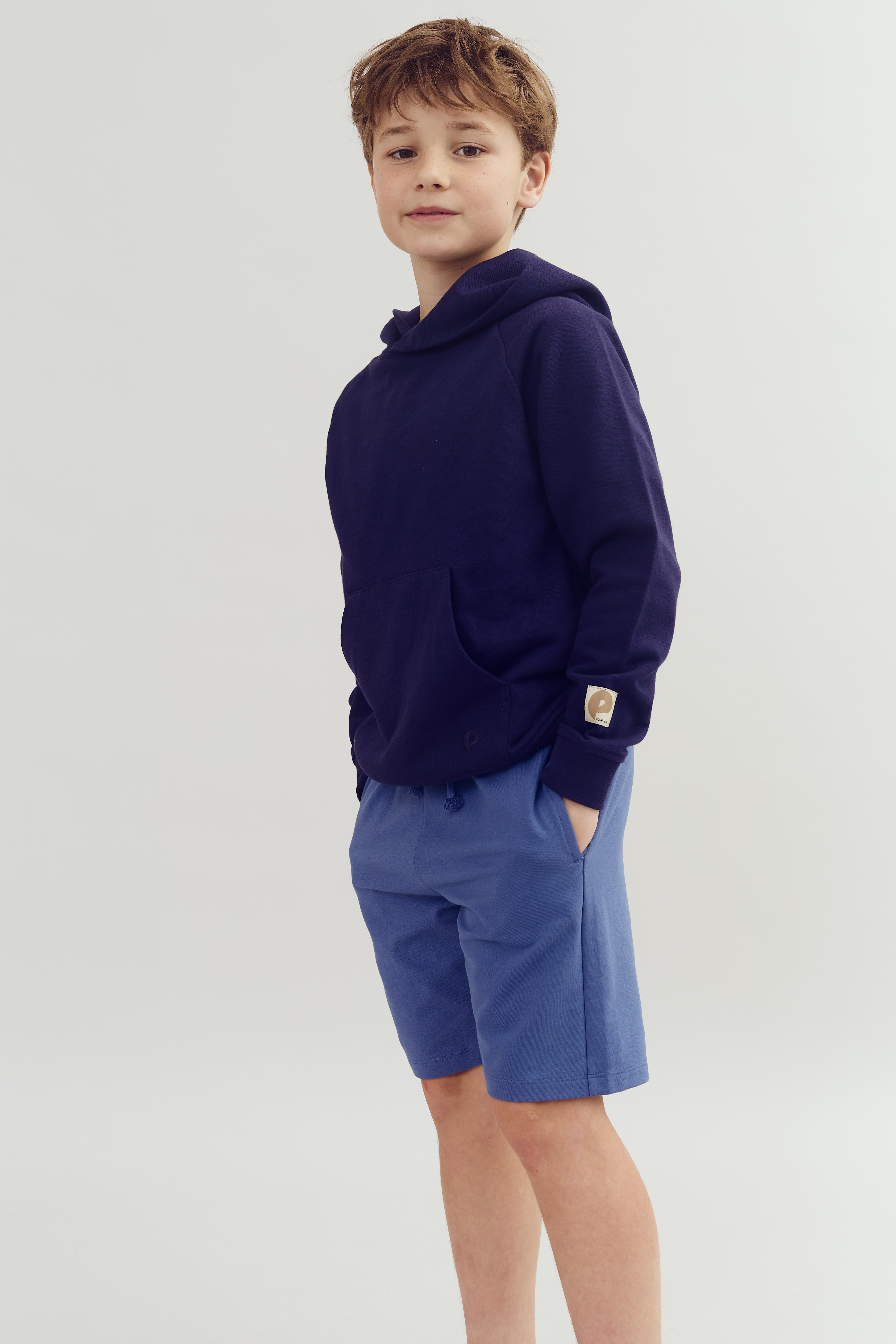 Ein Junge trägt einen dunkelblauen Hoodie und blaue Shorts, mit Händen in den Taschen, vor hellem Hintergrund. Der Hoodie hat ein kleines Logo am Ärmel.
