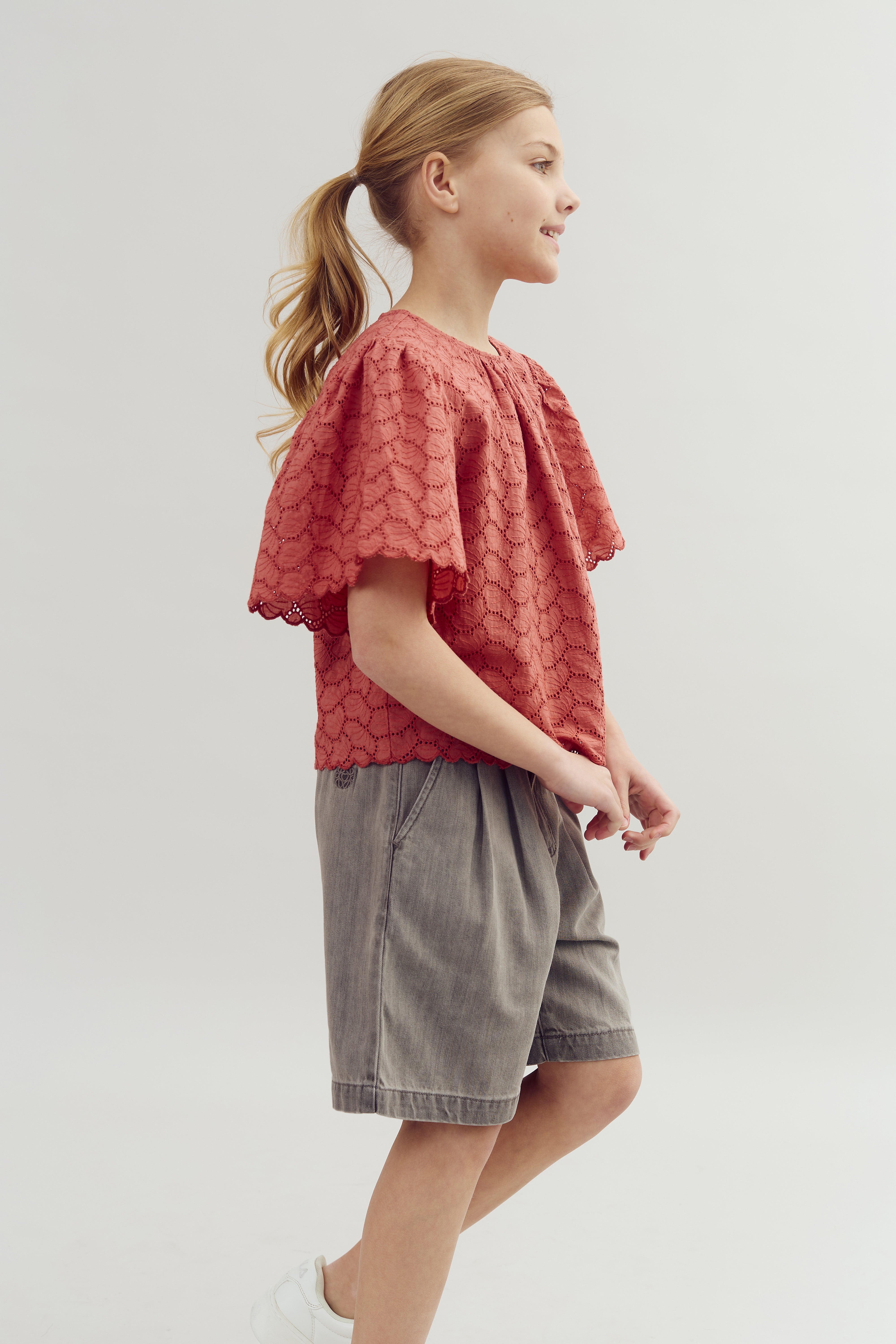 Ein Kind trägt ein rotes Spitzenoberteil und graue Shorts, geht lächelnd nach rechts. Der Hintergrund ist neutral und schlicht.