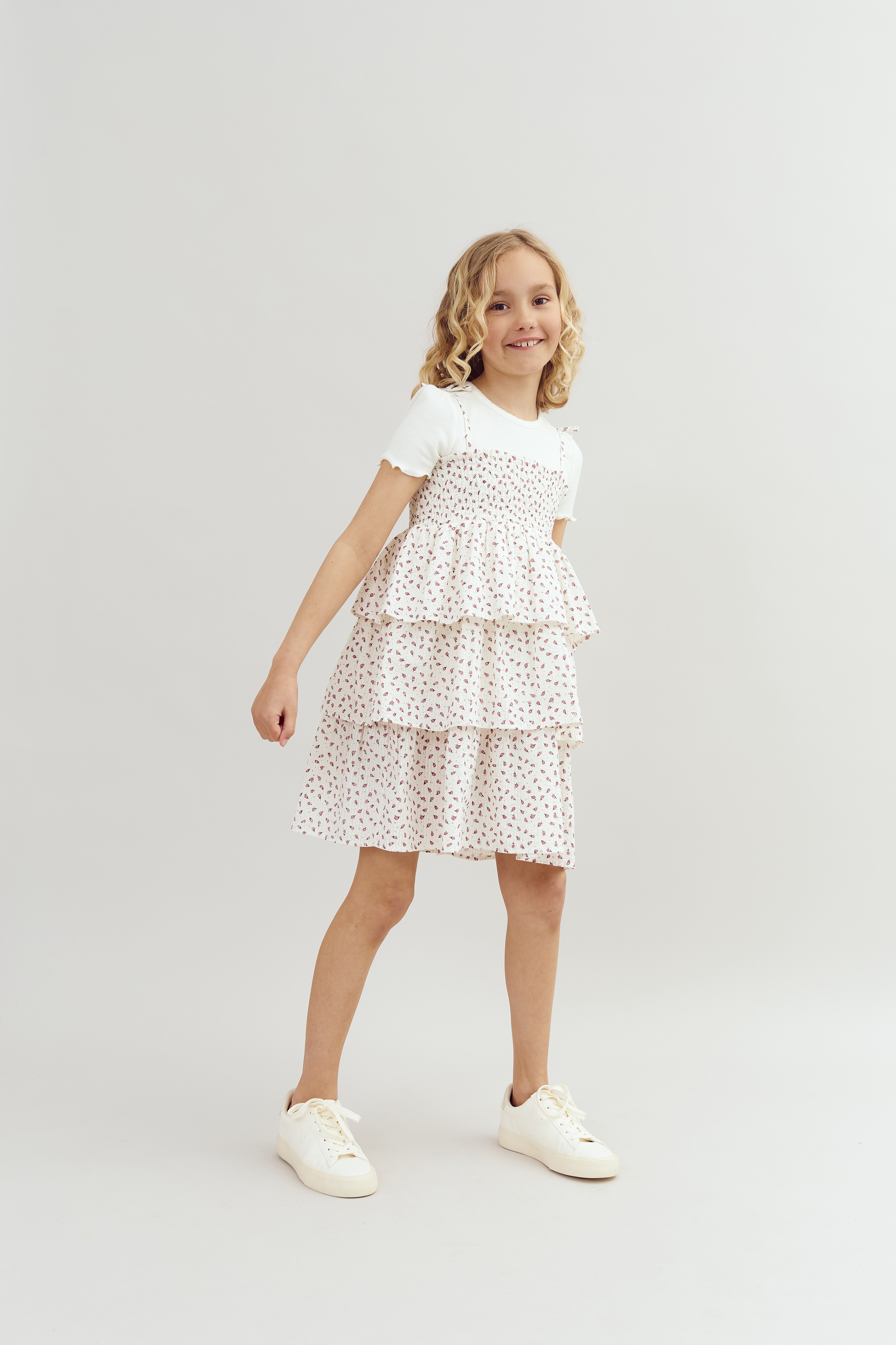 Ein Mädchen steht lächelnd in einem weißen, blumengemusterten Kleid mit Rüschen. Sie trägt weiße Turnschuhe. Der Hintergrund ist neutral und schlicht.